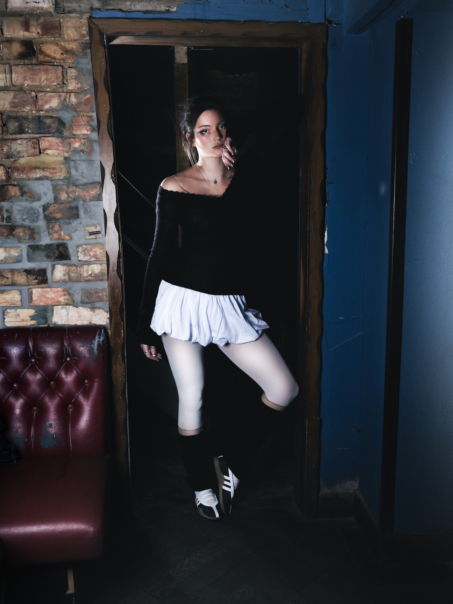 A woman wearing a fitted black top and a white skirt stands in a doorway. She has knee-high black socks and is posed with one finger to her lips, surrounded by a rustic interior featuring exposed brick and a red leather couch.