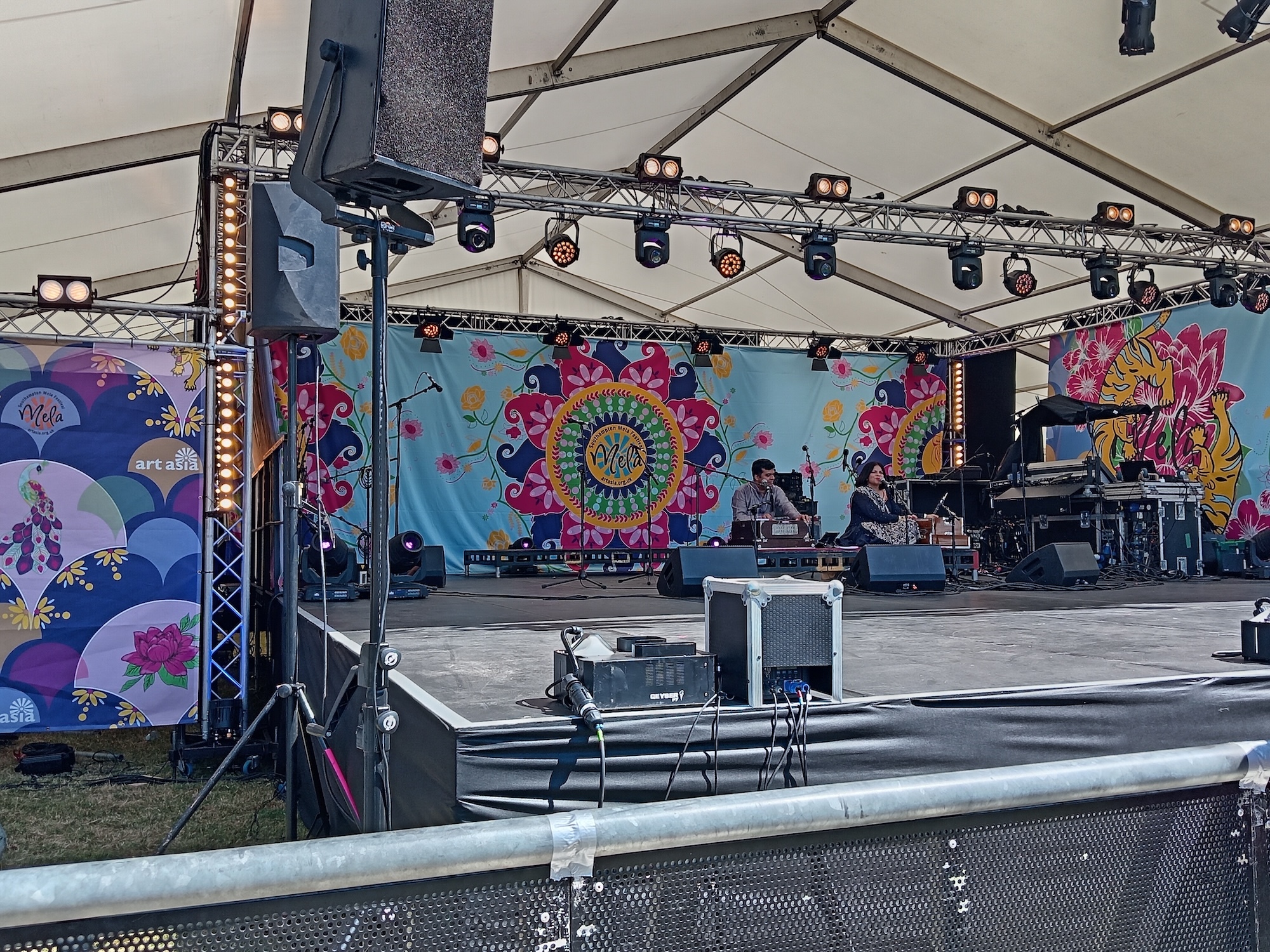 Outdoor stage with white canopy, metal truss lighting rig, and colourful mandala-style backdrop featuring blues, pinks, and yellows.