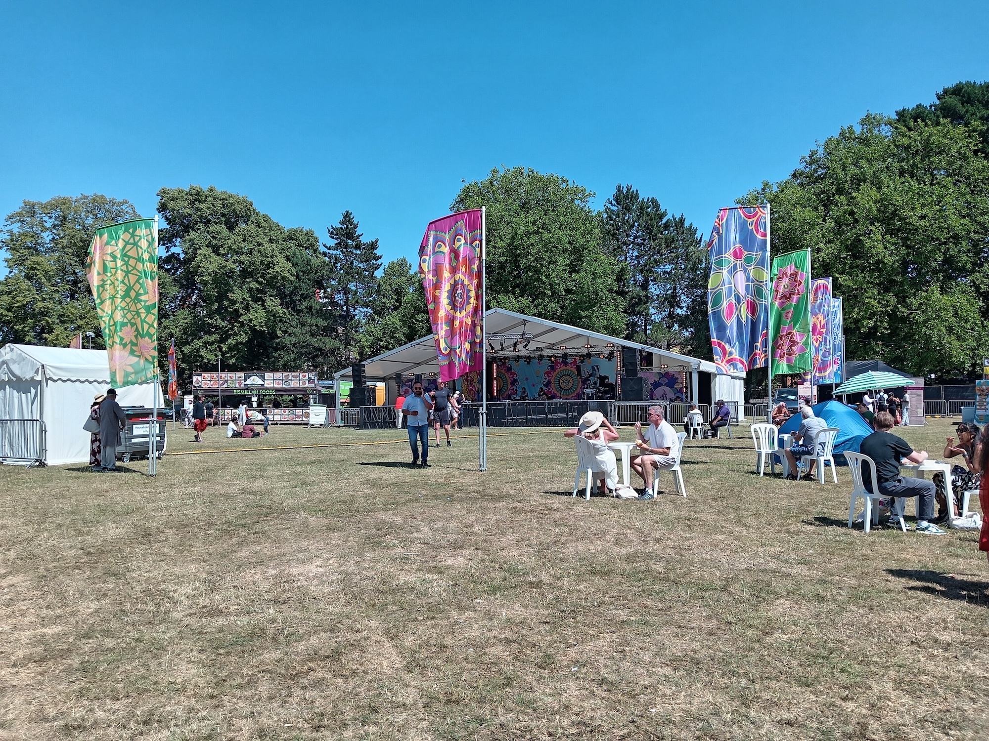 Outdoor festival with colourful banners, white tent, people seated at tables on grass field, trees and blue sky in background.