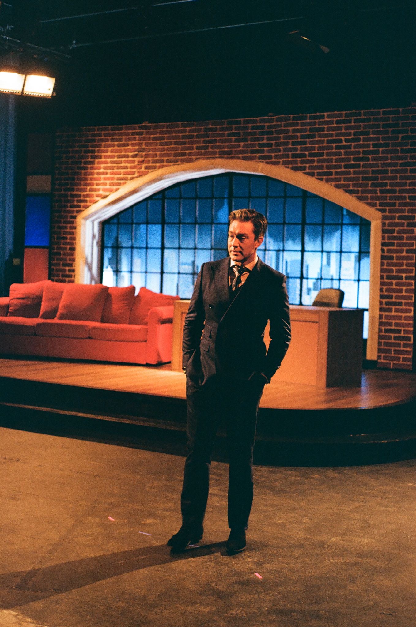 A man in a black suit stands on a television studio set, with a brick backdrop and a blue window design. A red sofa is positioned in front of a wooden desk, indicating a talk show setting.