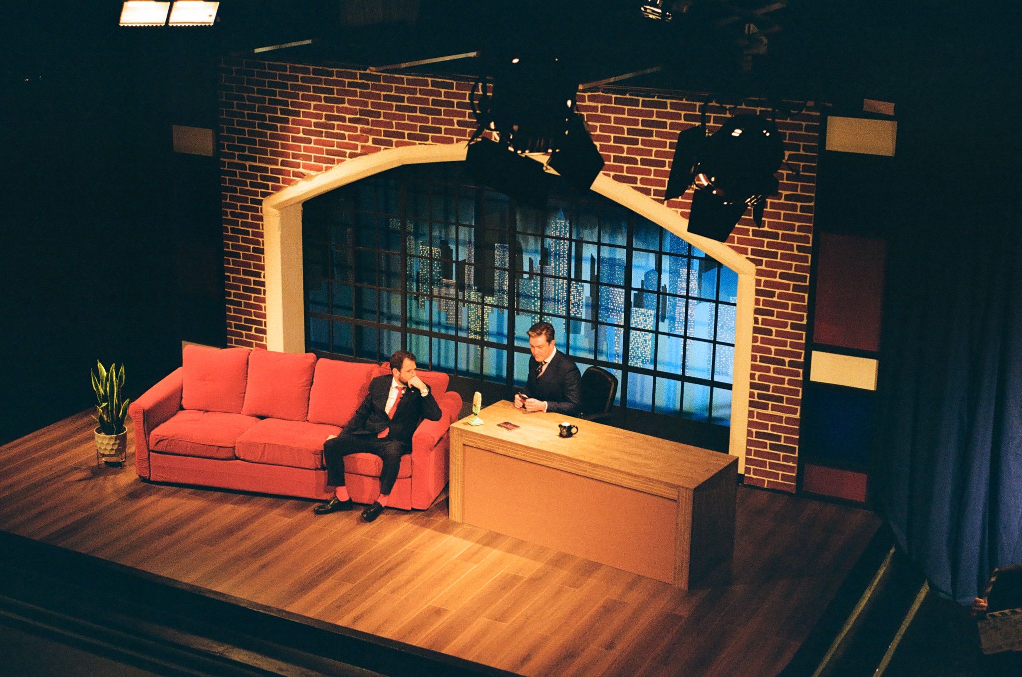 A television studio set featuring a red sofa, where a man in a black suit sits, and another man in a white shirt is seated at a desk. The background has large windows with a blue-lit backdrop and a potted plant beside the sofa.