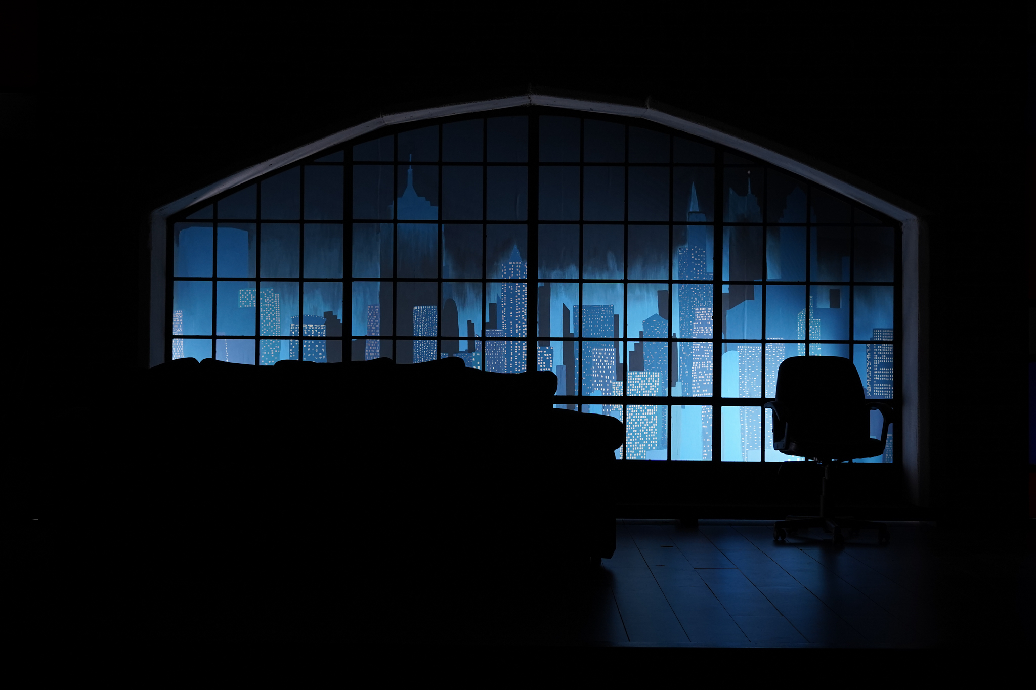 A dark room featuring a large window with a silhouette of a city skyline illuminated in shades of blue. In the foreground, a dark couch is visible, along with a black chair positioned beside it, against a wooden floor.