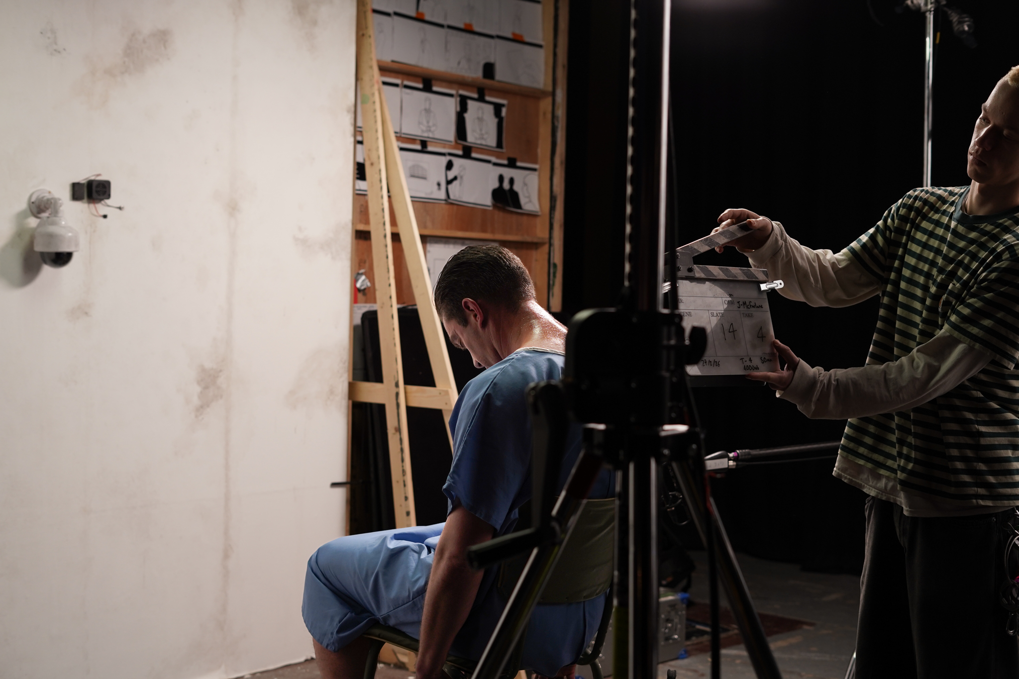 A man in a blue medical scrub sits on a chair, facing away from the camera. A second person, dressed in a striped shirt, is holding a clapperboard in front of him. The background features a bare wall with shelves and lighting equipment.