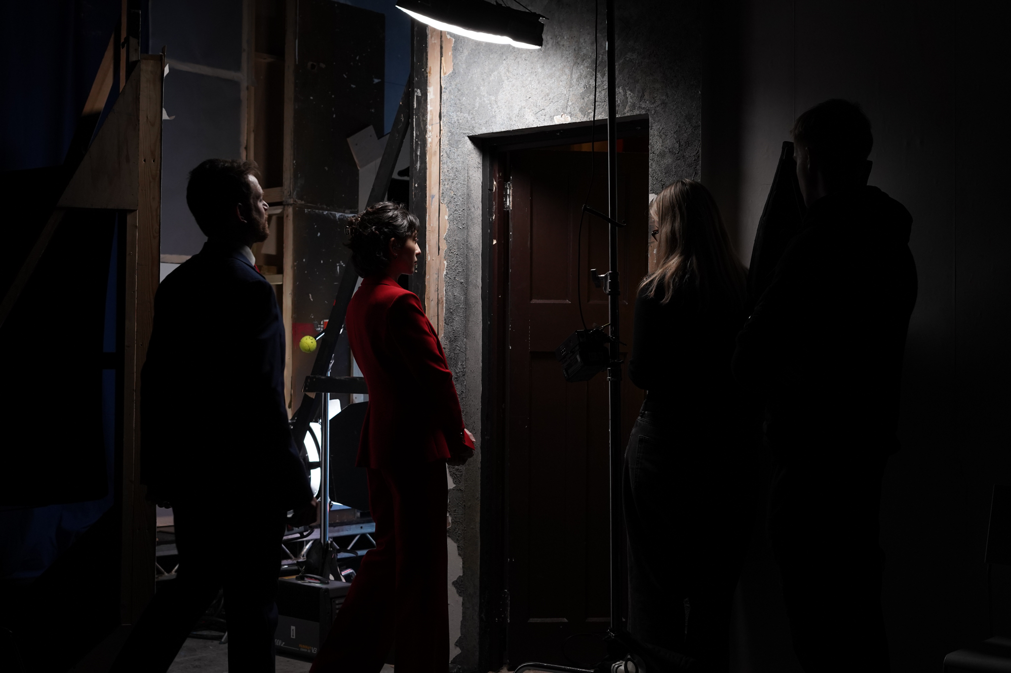 A woman in a red suit stands facing a closed door, illuminated by a light above. Three others, including a man in a dark jacket and a woman in a light-coloured outfit, are positioned nearby in a dimly lit room with visible filmmaking equipment.