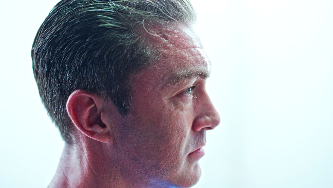 A close-up side profile of a man with dark, slicked-back hair and a serious expression. His skin appears slightly illuminated against a soft, bright background, highlighting his features.