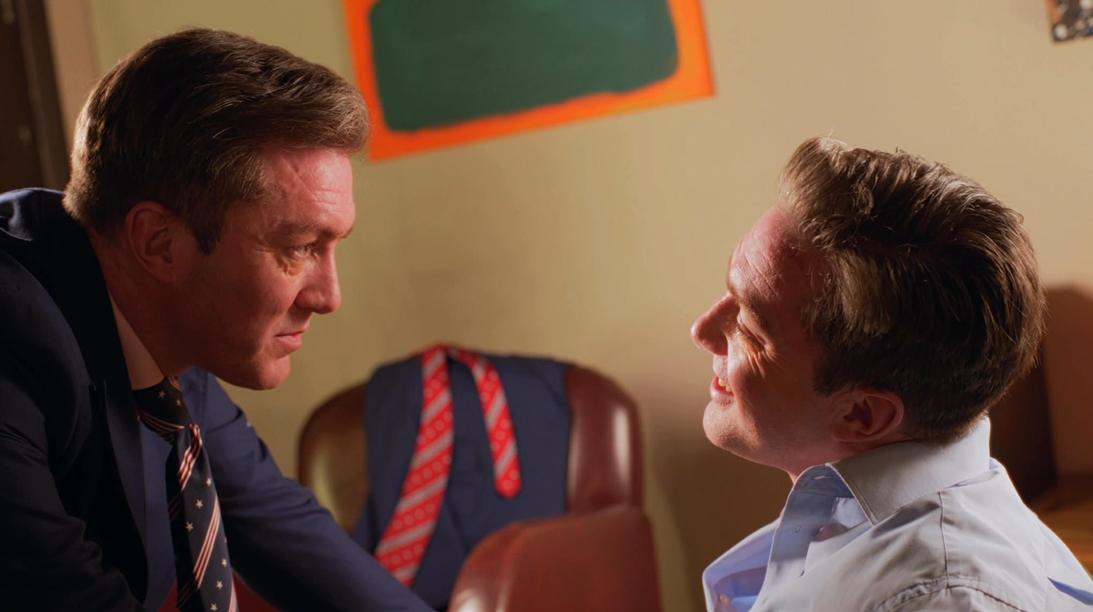 Two men facing each other in a close-up shot, one dressed in a dark suit with a tie and the other in a light blue shirt. A chalkboard and an empty chair are visible in the background, along with a jacket hanging on a chair.
