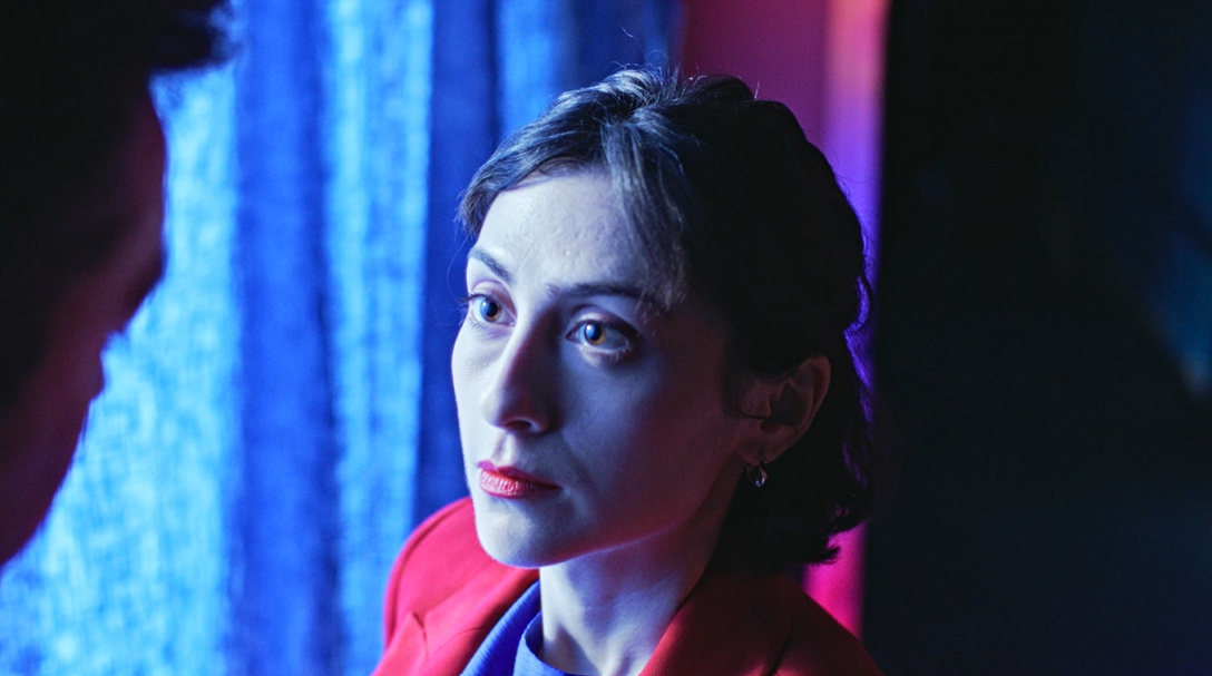 A woman with shoulder-length dark hair is positioned in front of a blue background with soft lighting. She wears a bright red blazer over a purple top, and her gaze is directed towards the viewer, who is partially out of frame.