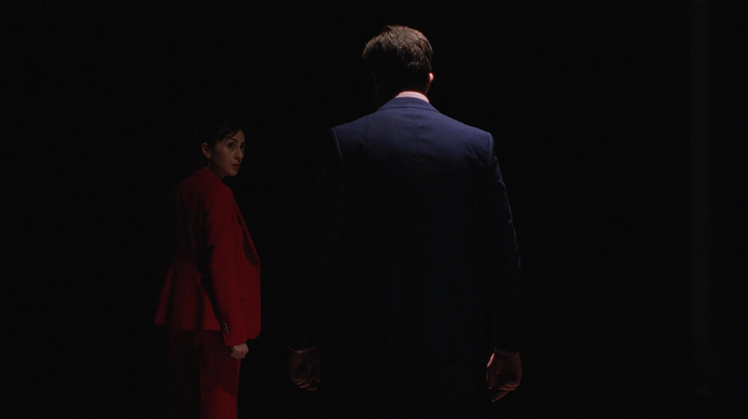 A woman dressed in a red suit stands facing slightly to the side, looking back at a man in a dark suit. They are both illuminated by soft light against a dark background.
