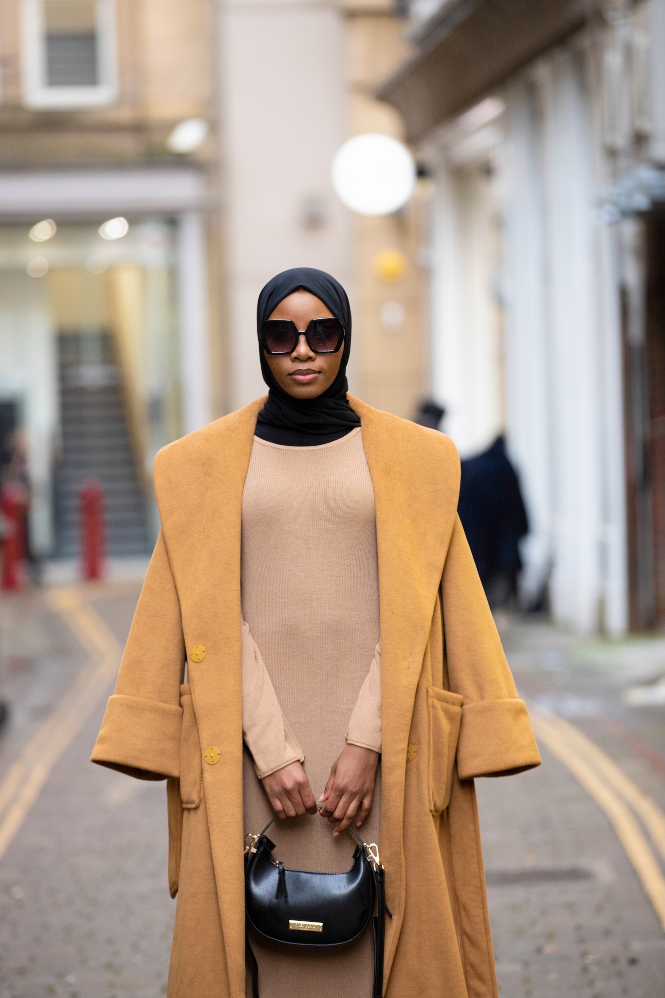 Woman wearing black hijab, sunglasses, beige dress and mustard yellow coat, holding black handbag on urban street.