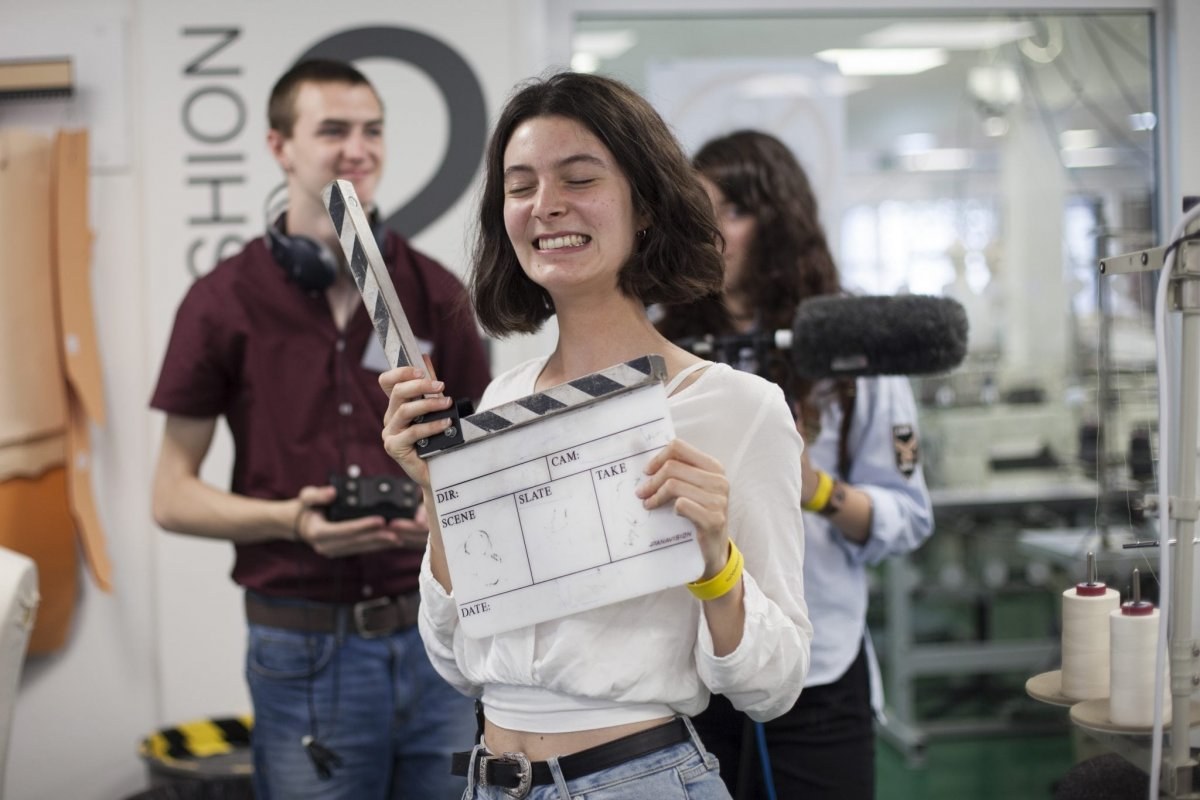 Person holding a clapper board with their eyes closed. Two other people are stood behind them in the background.