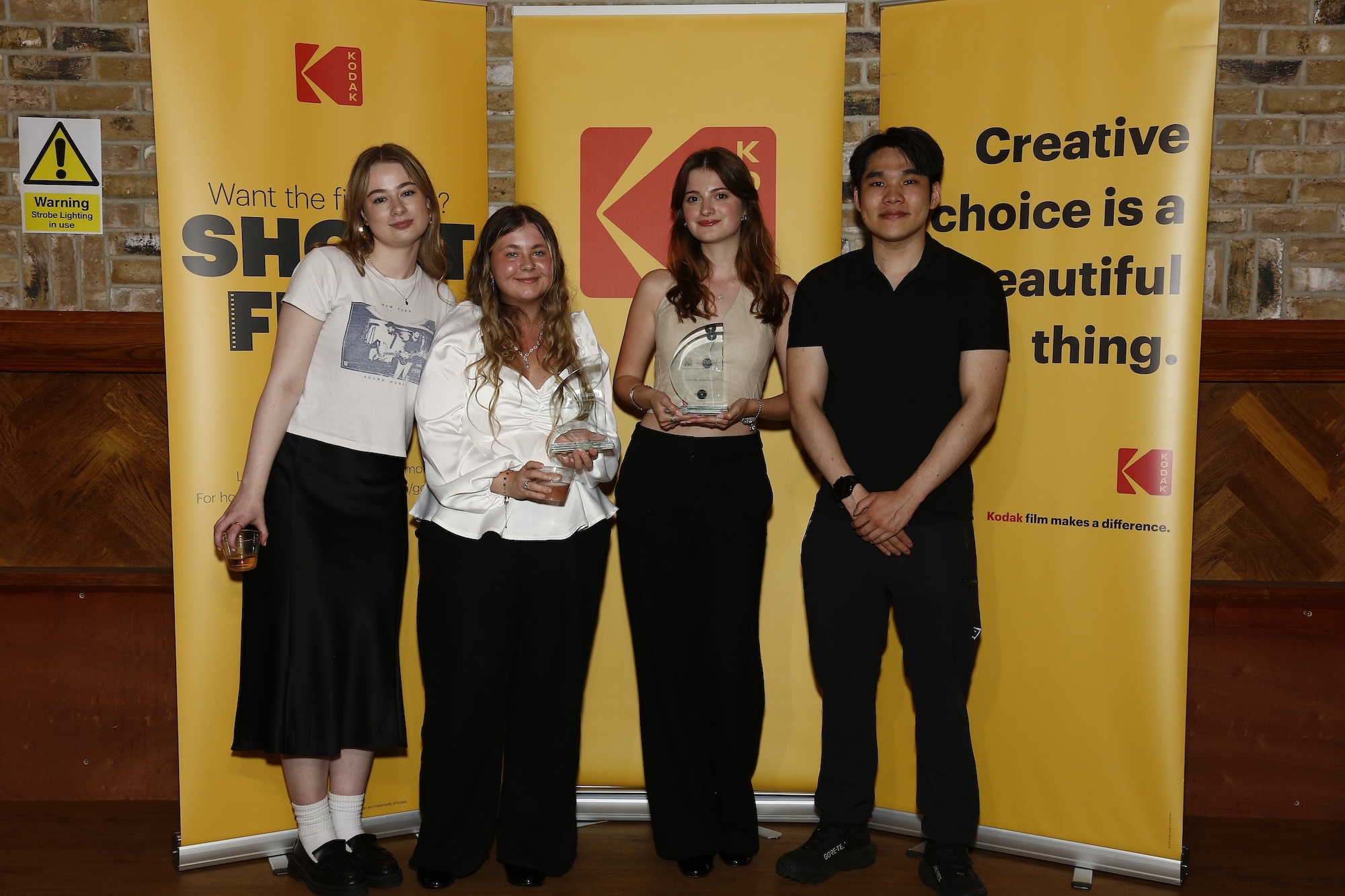 Four Arts University Bournemouth BA (Hons) Film Production students holding two awards won at the Kodak/NAHEMI Student Commercial Awards, posing for a photo in front of three yellow banners.