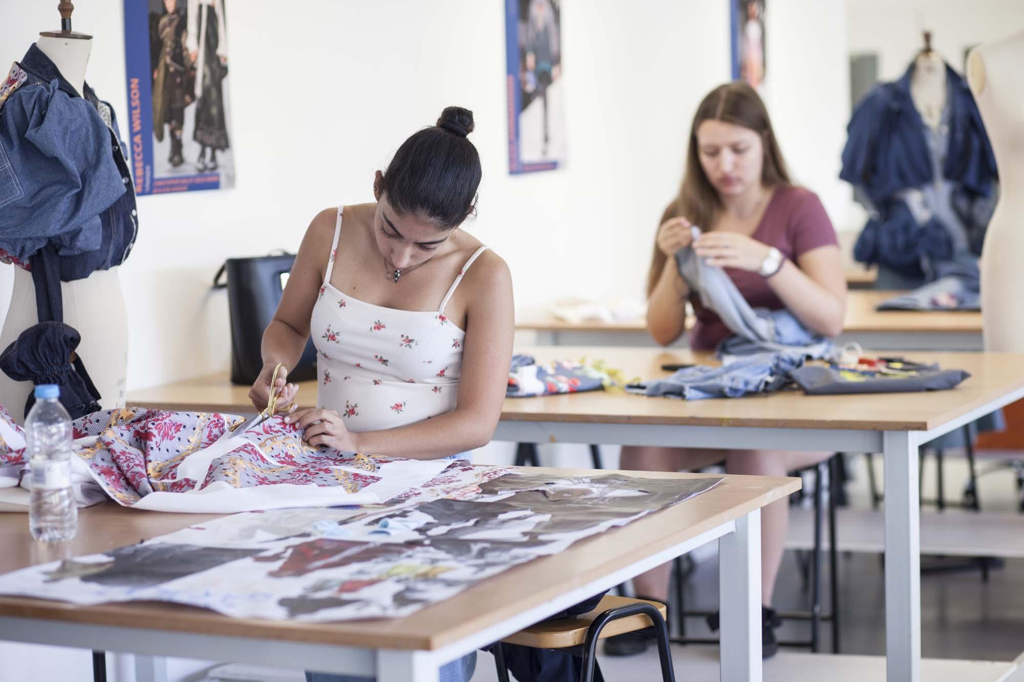 Two students sitting at tables in a costume studio working on different pieces of fabric.