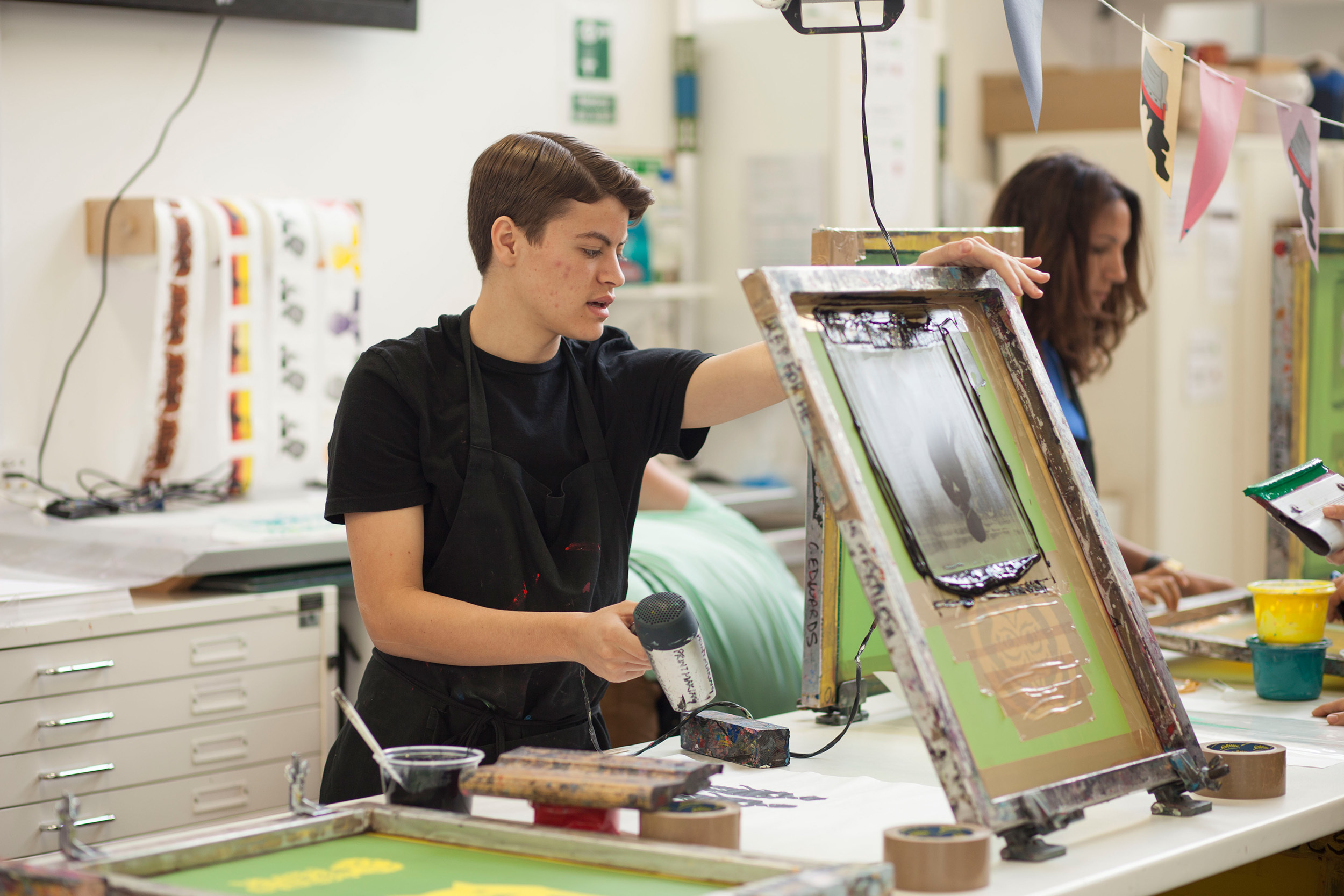 Students in the print room at AUB, one student has just finished screenprinting and is drying the artwork using a dryer