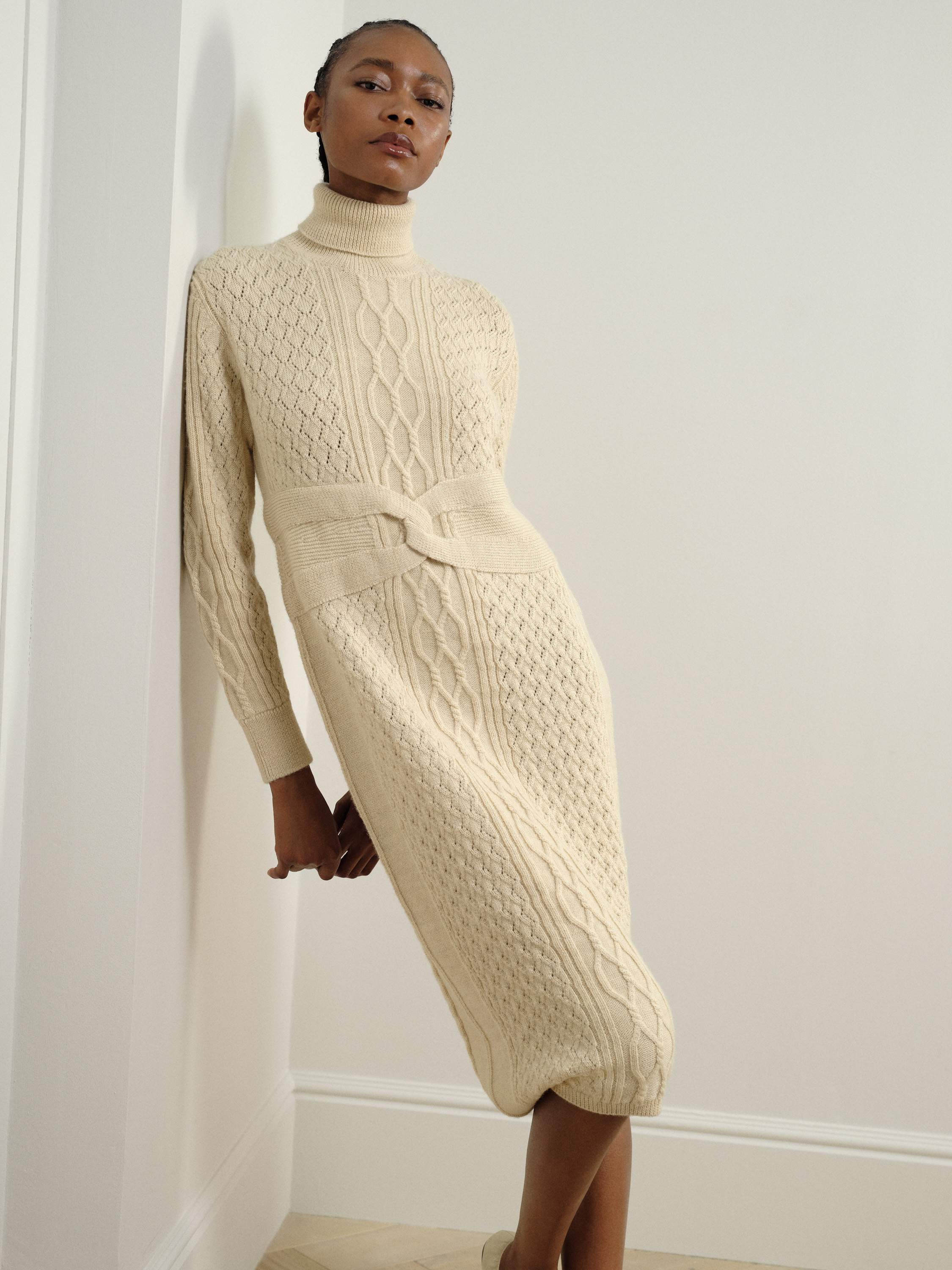 A model poses against a wall while wearing a cream-coloured woollen garment.