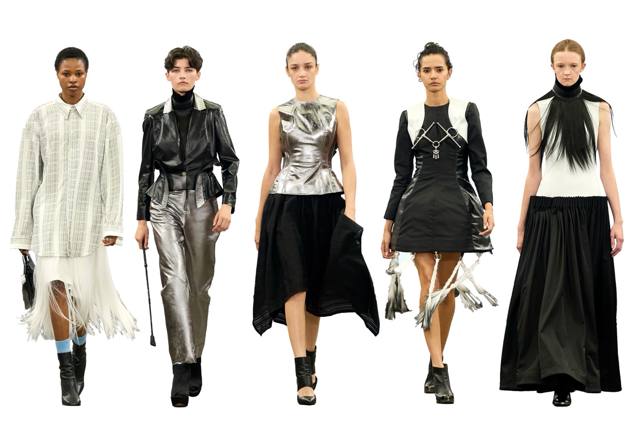 Five fashion models walk a runway in avant-garde black, white and metallic outfits against a plain white background.