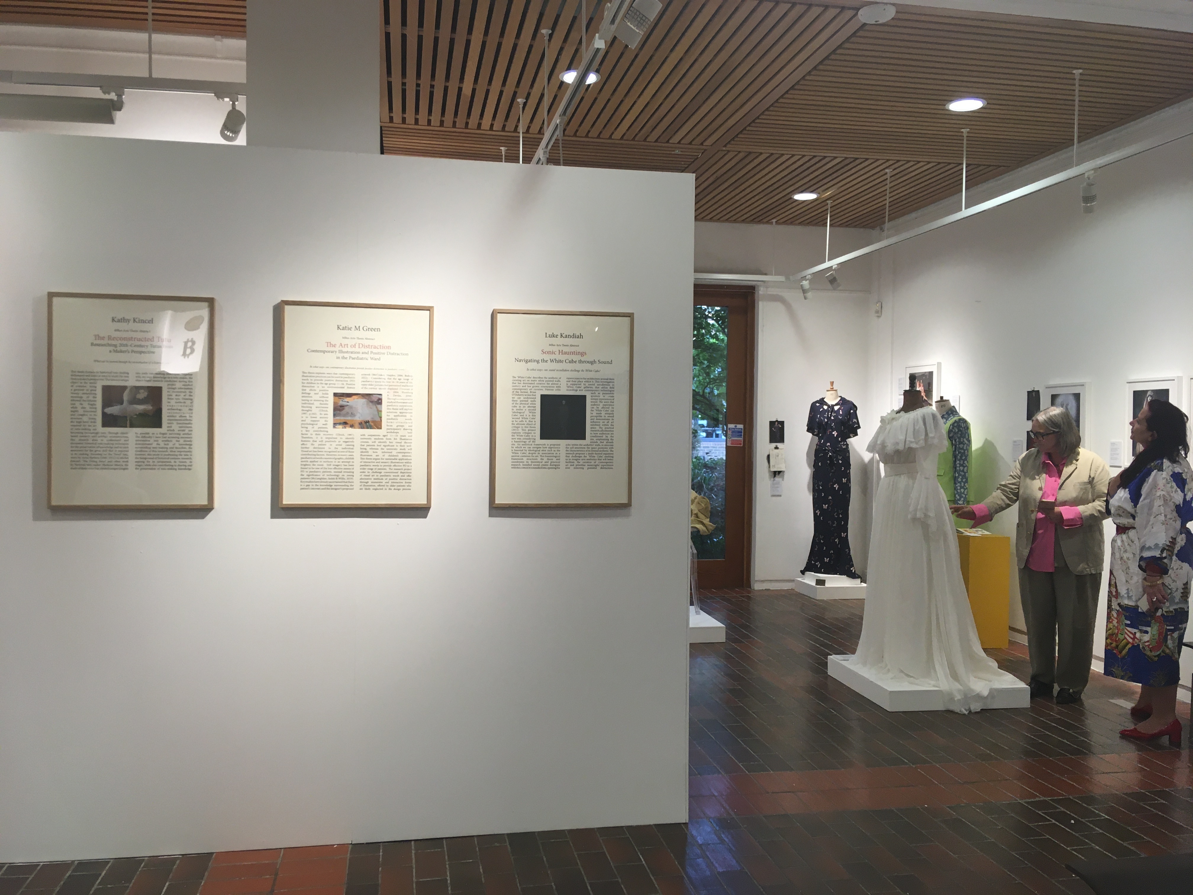 Three pages each with a body of text and a picture in the centre in frames on a gallery wall. Two people are standing to the far right next to mannequins dressed with costumes.
