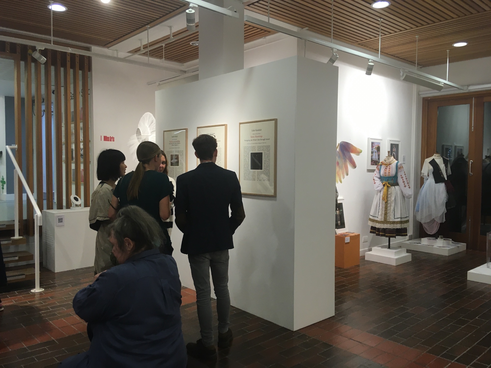 A small group of people look at three framed pieces of artwork on a gallery wall.