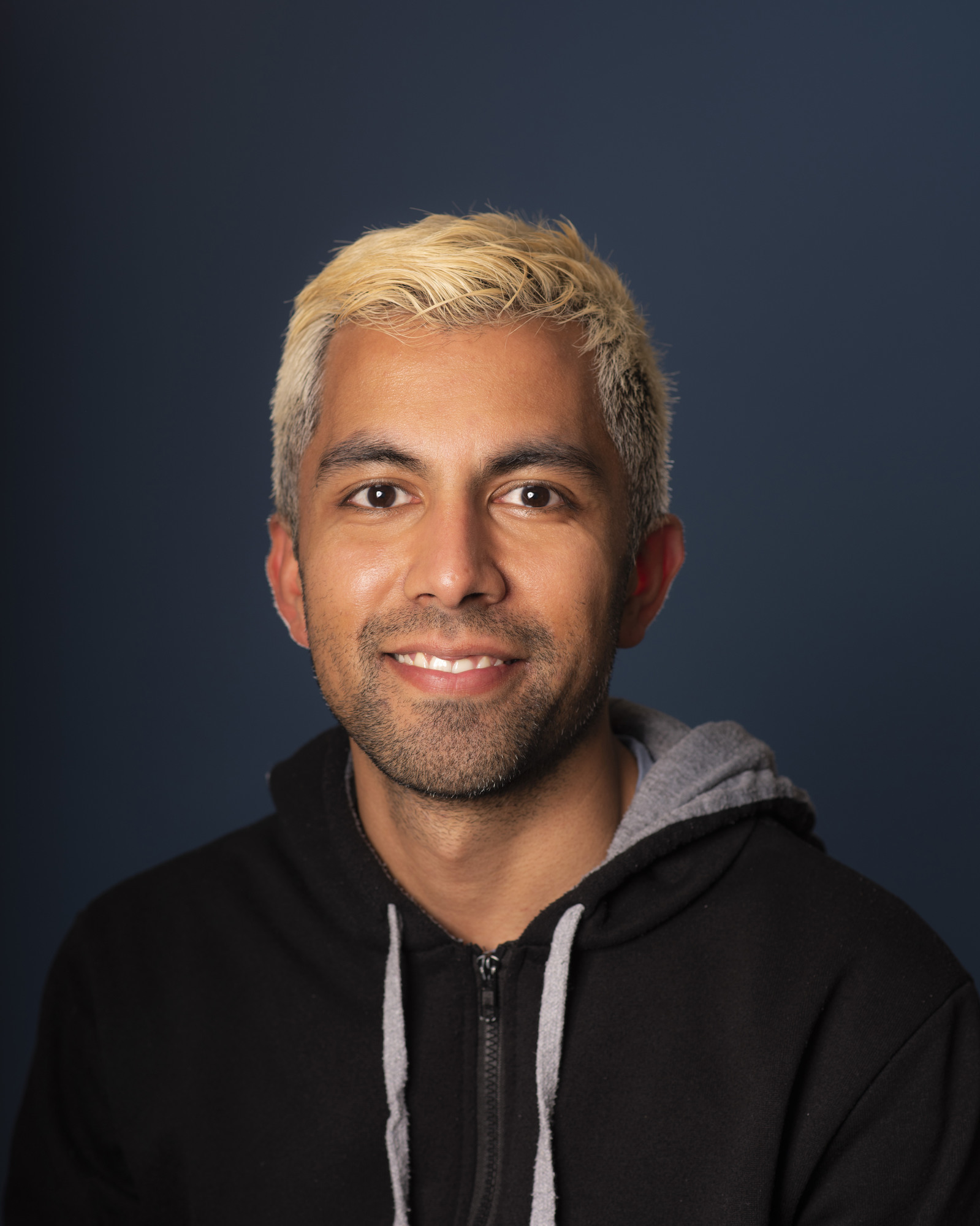 Man with bleached blonde hair and facial hair wearing black hoodie with grey hood, smiling against dark blue background.