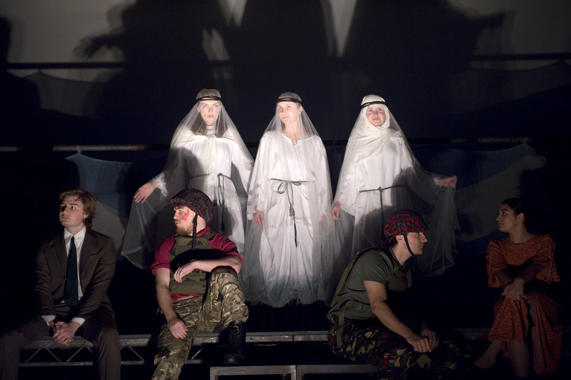 A group of BA (Hons) Acting students performing Macbeth. The spotlight is focused on three students in white robes portraying the Witches, with other performers sat in front of them.