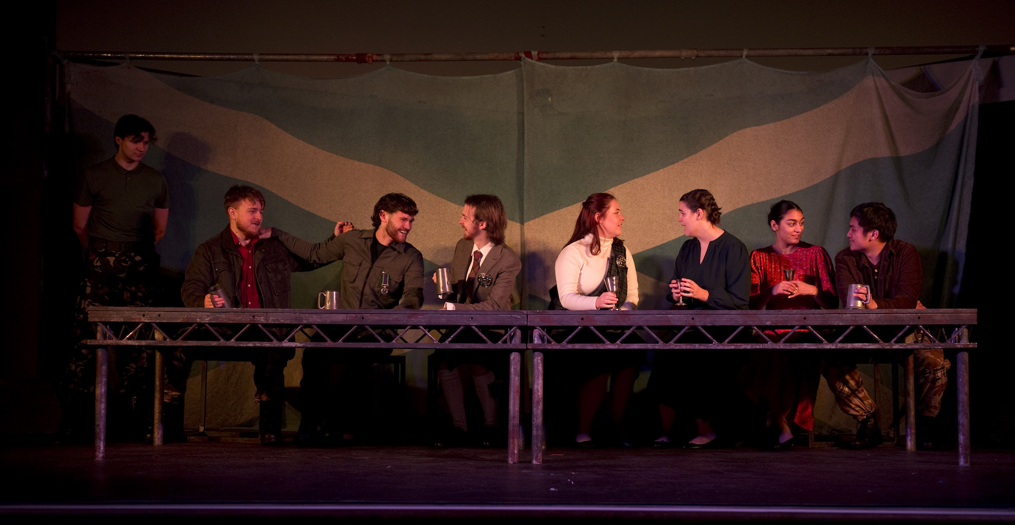 A group of BA (Hons) Acting students sitting on stage performing Macbeth. They are all sat along a long metal frame, made to look like a table. A large Scottish flag hangs in the background.