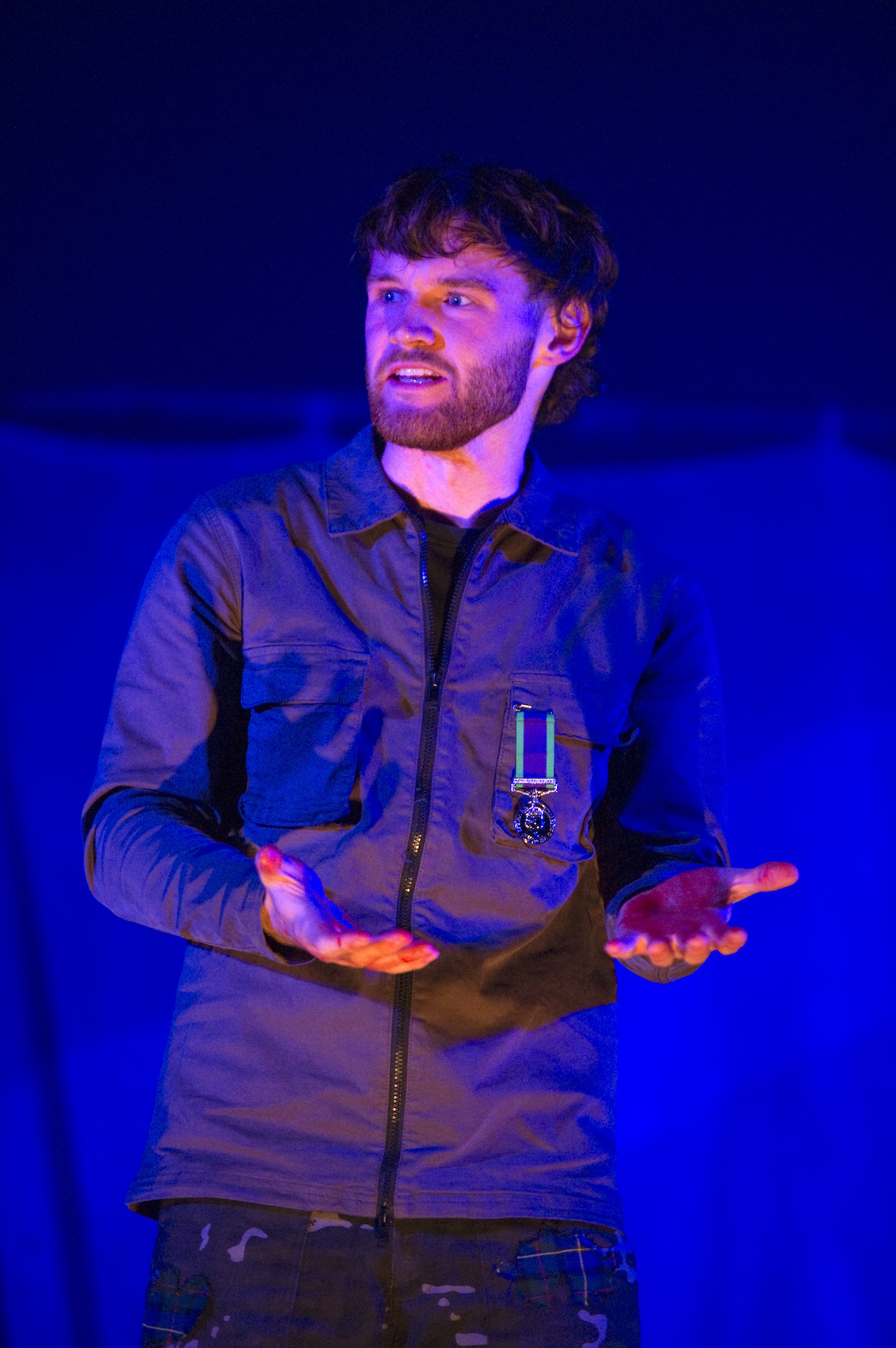 A BA (Hons) Acting student performing Macbeth on stage with a blue light behind, their hands are held out and have blood on them.