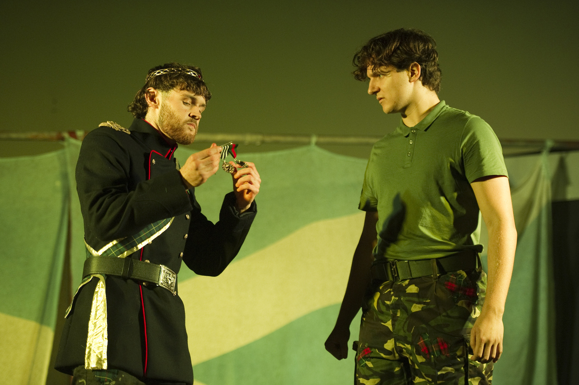 Two BA (Hons) Acting students on stage at the Palace Court Theatre performing Macbeth. One actor in a military tunic and crown handles a medal while another actor in military fatigues watches.
