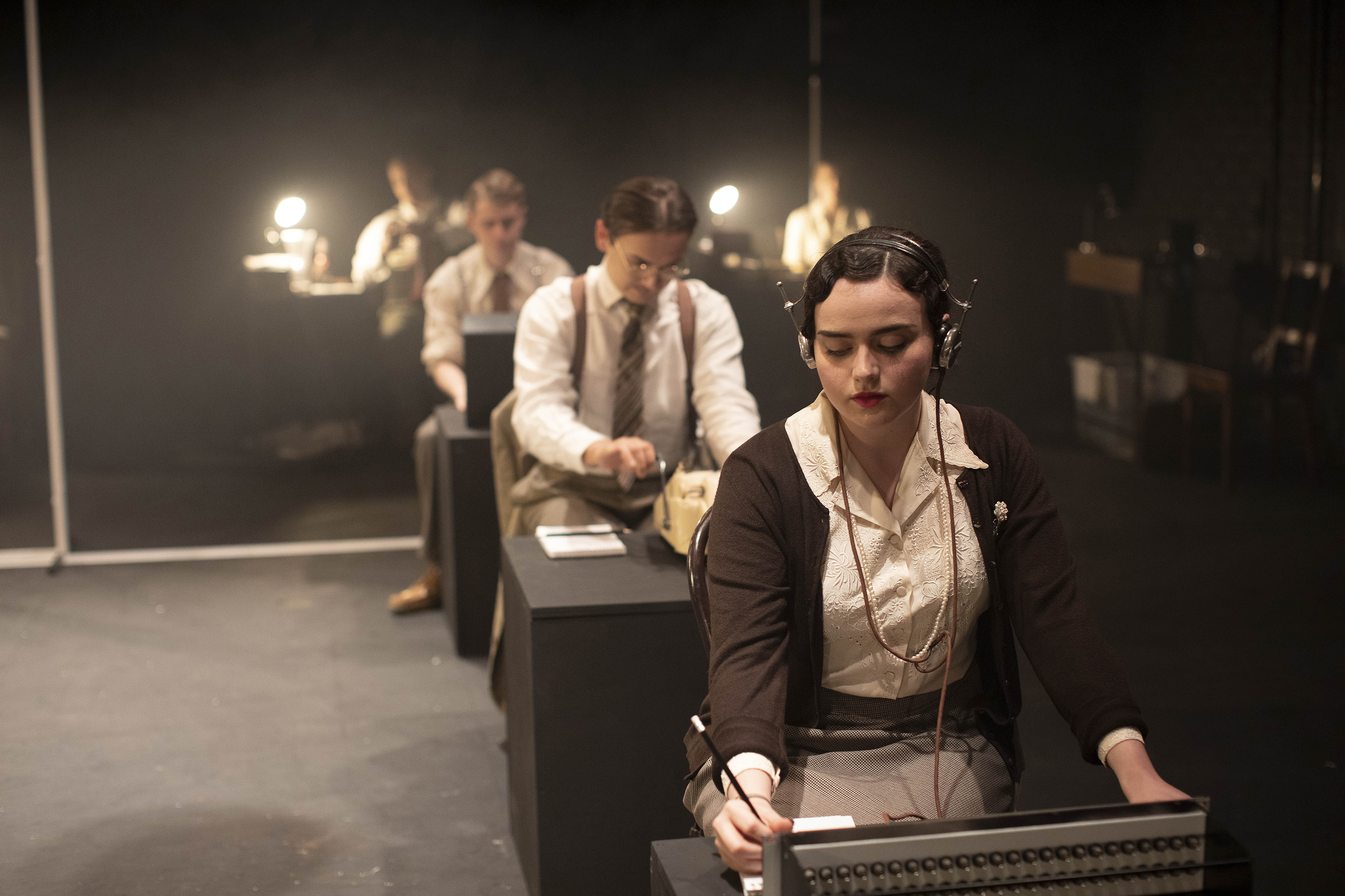 Row of people in period costumes sitting at tables. One person at the front is operating a switchboard wearing headphones.
