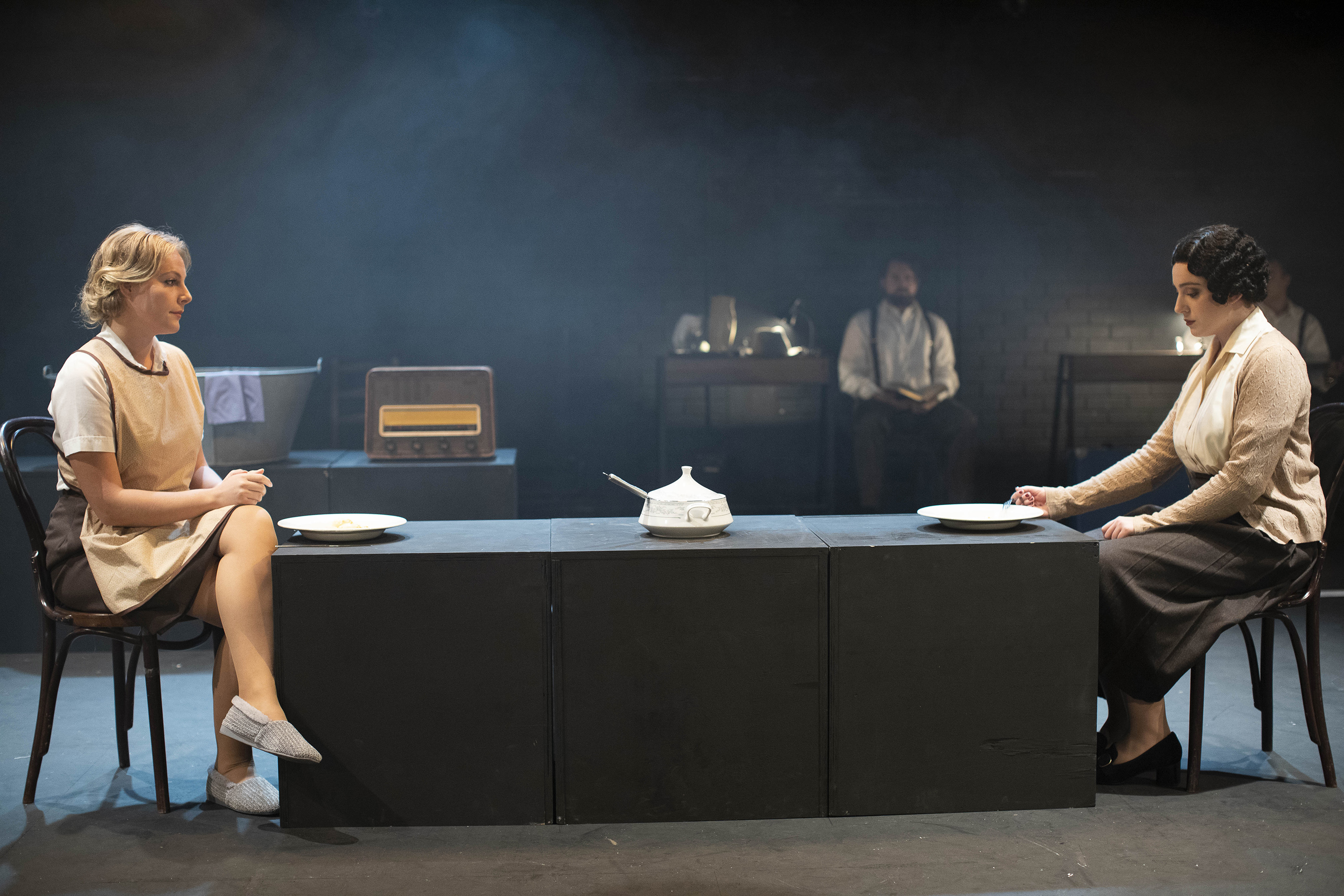 Two people in costume sit at opposite ends of a long table. There are soup bowls in front of them and a tureen in the middle of the table.