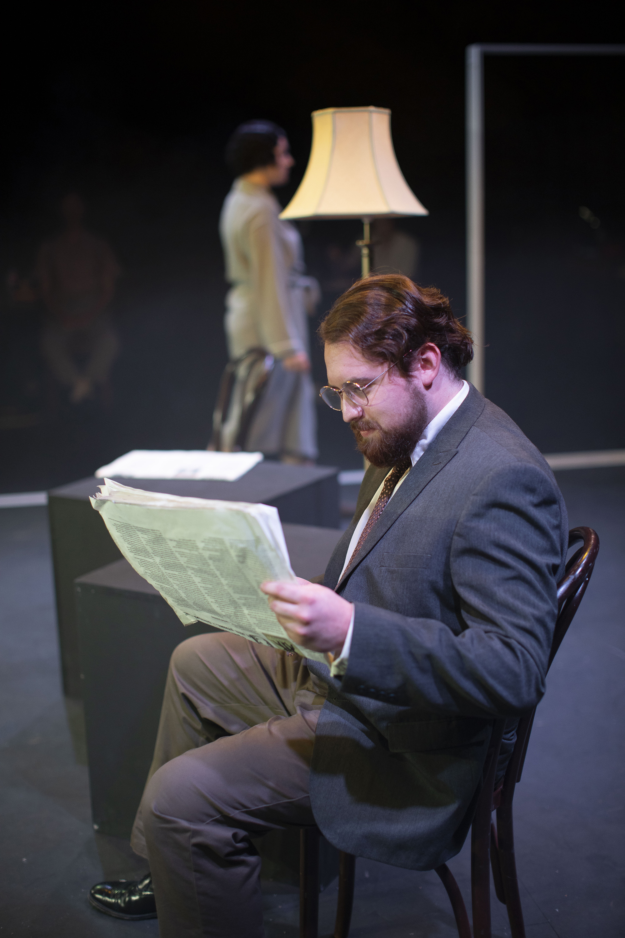 Performer in costume on stage sitting in a chair and reading a newspaper.