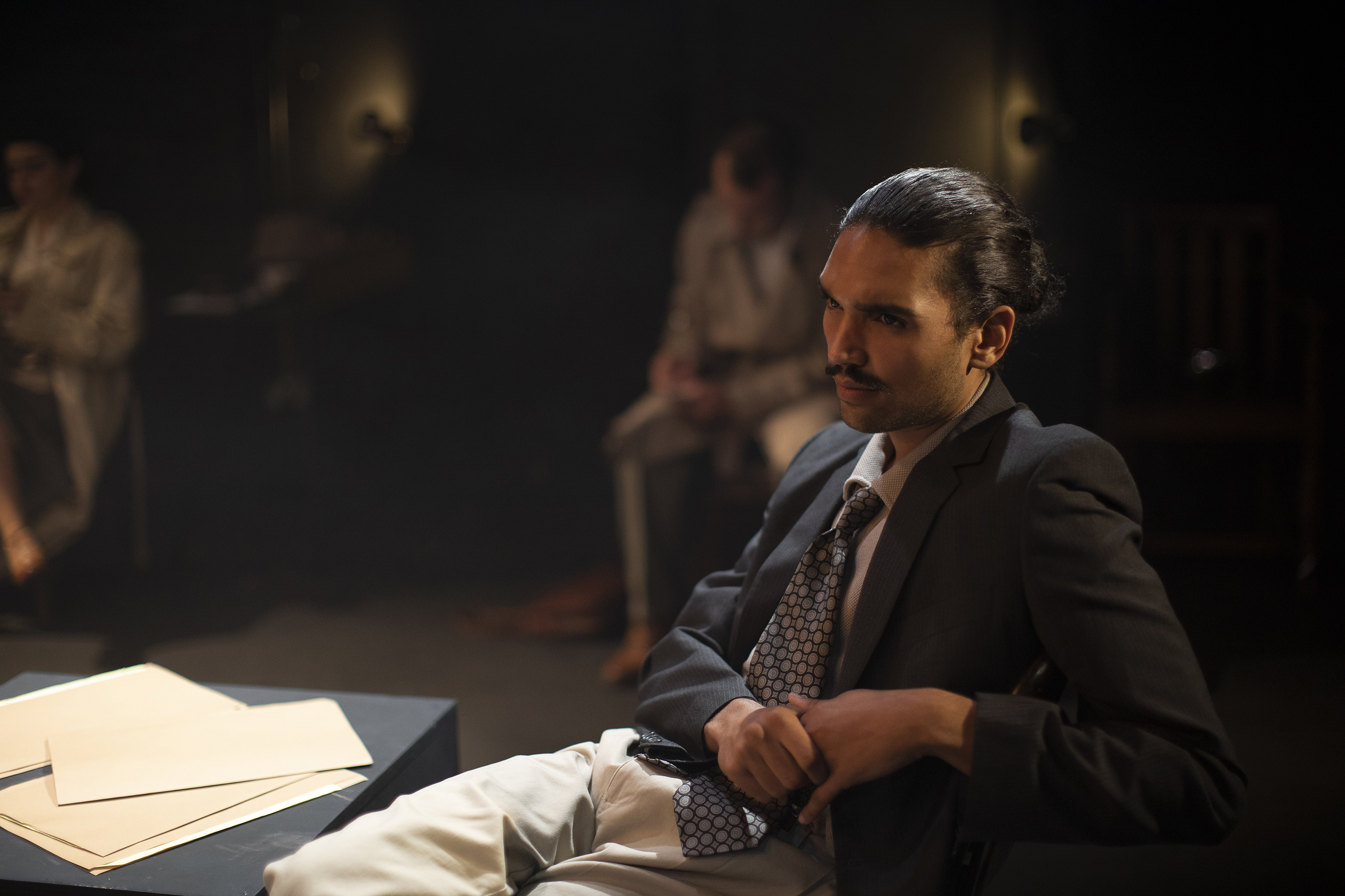 Person on stage sitting around in a chair next to a low table with papers strewn across it.