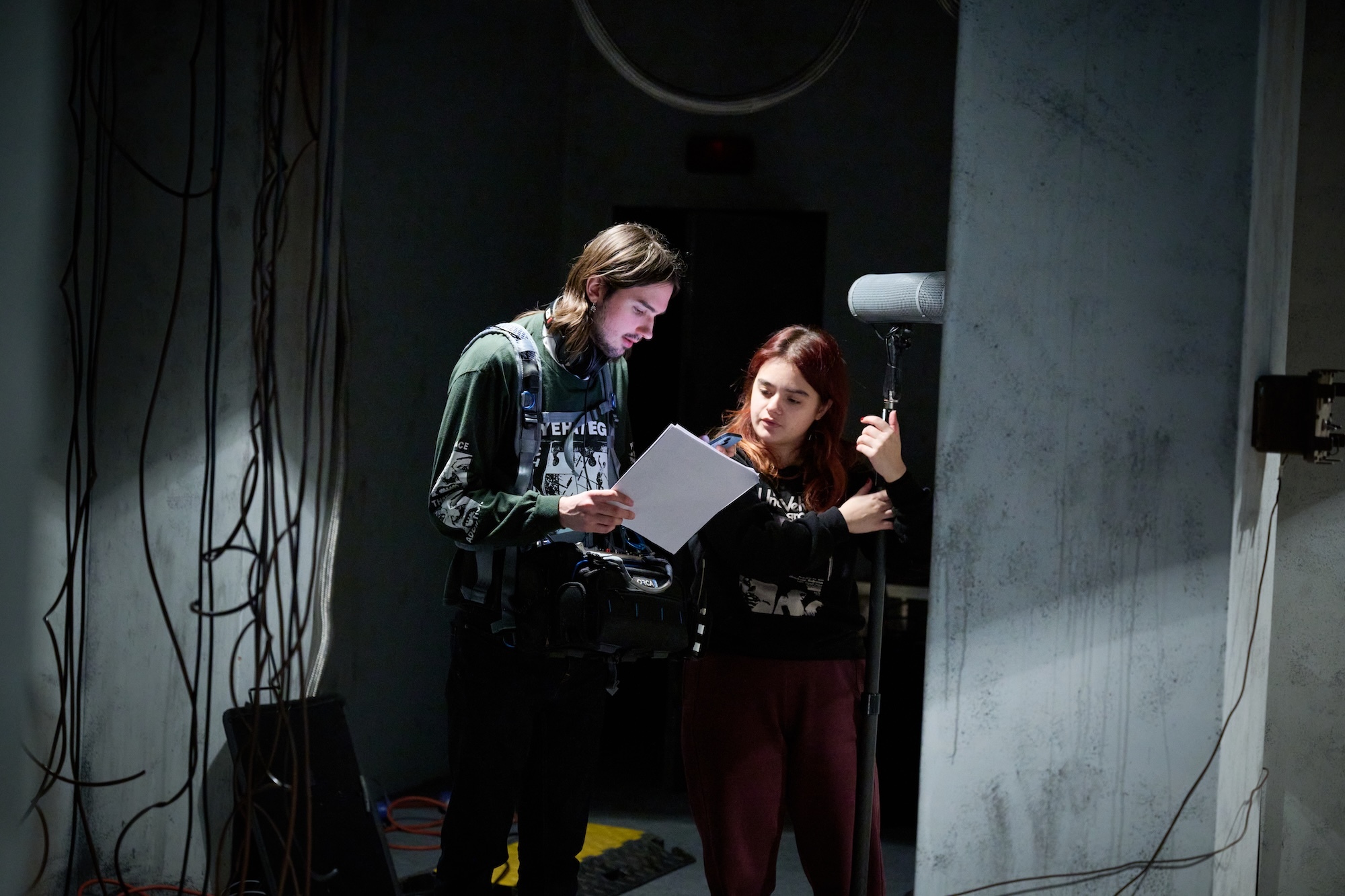Two people examining papers in dimly lit room with exposed cables and industrial equipment visible in background.