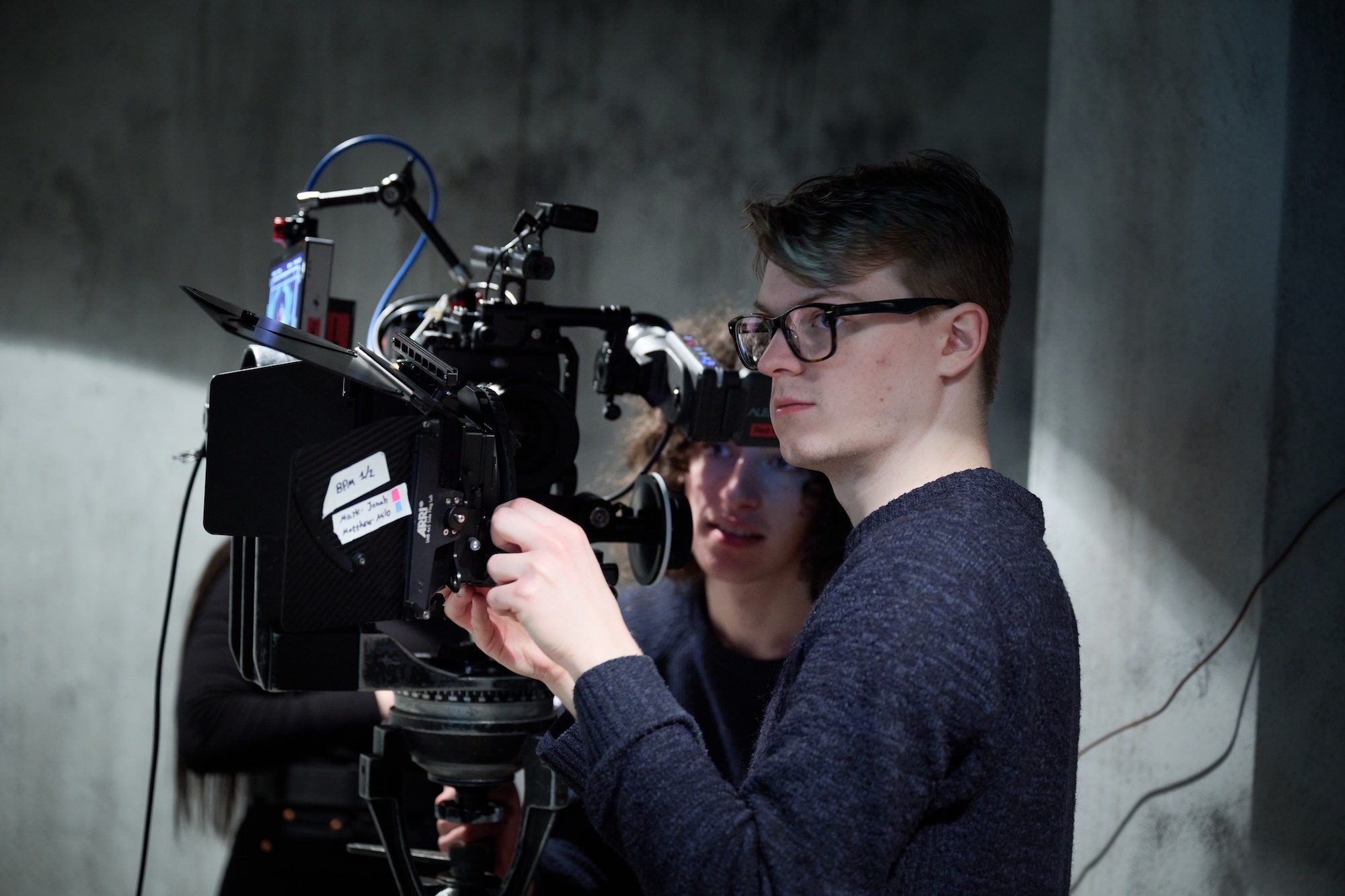 Man in glasses operates professional film camera with another person behind him in dimly lit indoor setting.