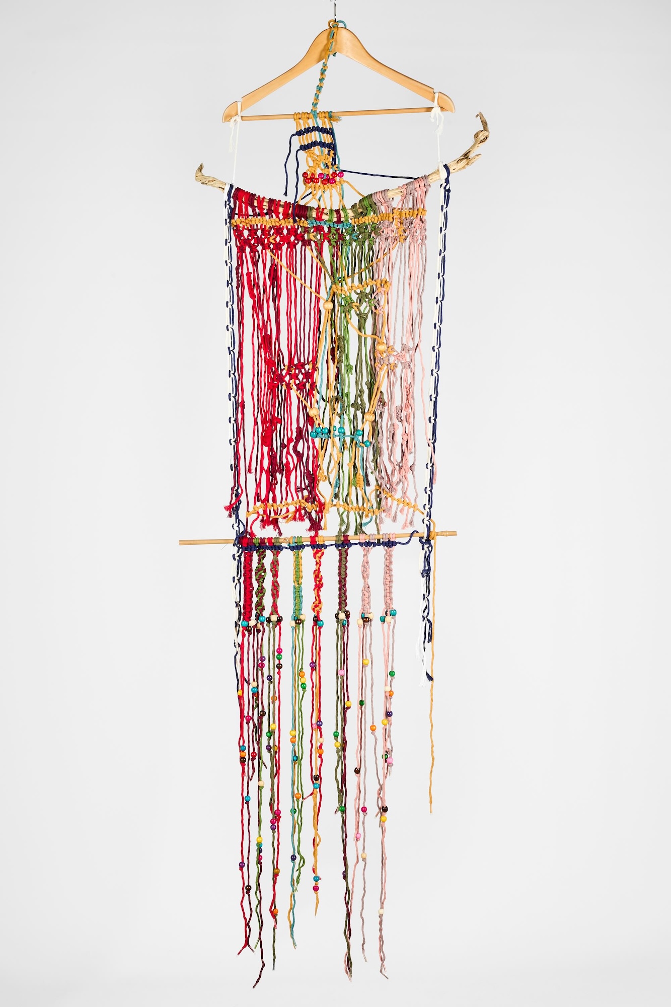 A colourful hanging textile piece with long braided strands and beads, suspended from a wooden hanger against a plain white background.