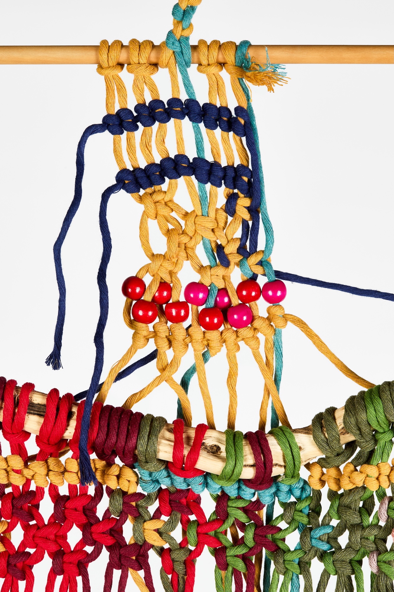 A close-up of a colourful woven macramé wall hanging with blue, yellow, red and green cords, supported by a wooden dowel against a white background.