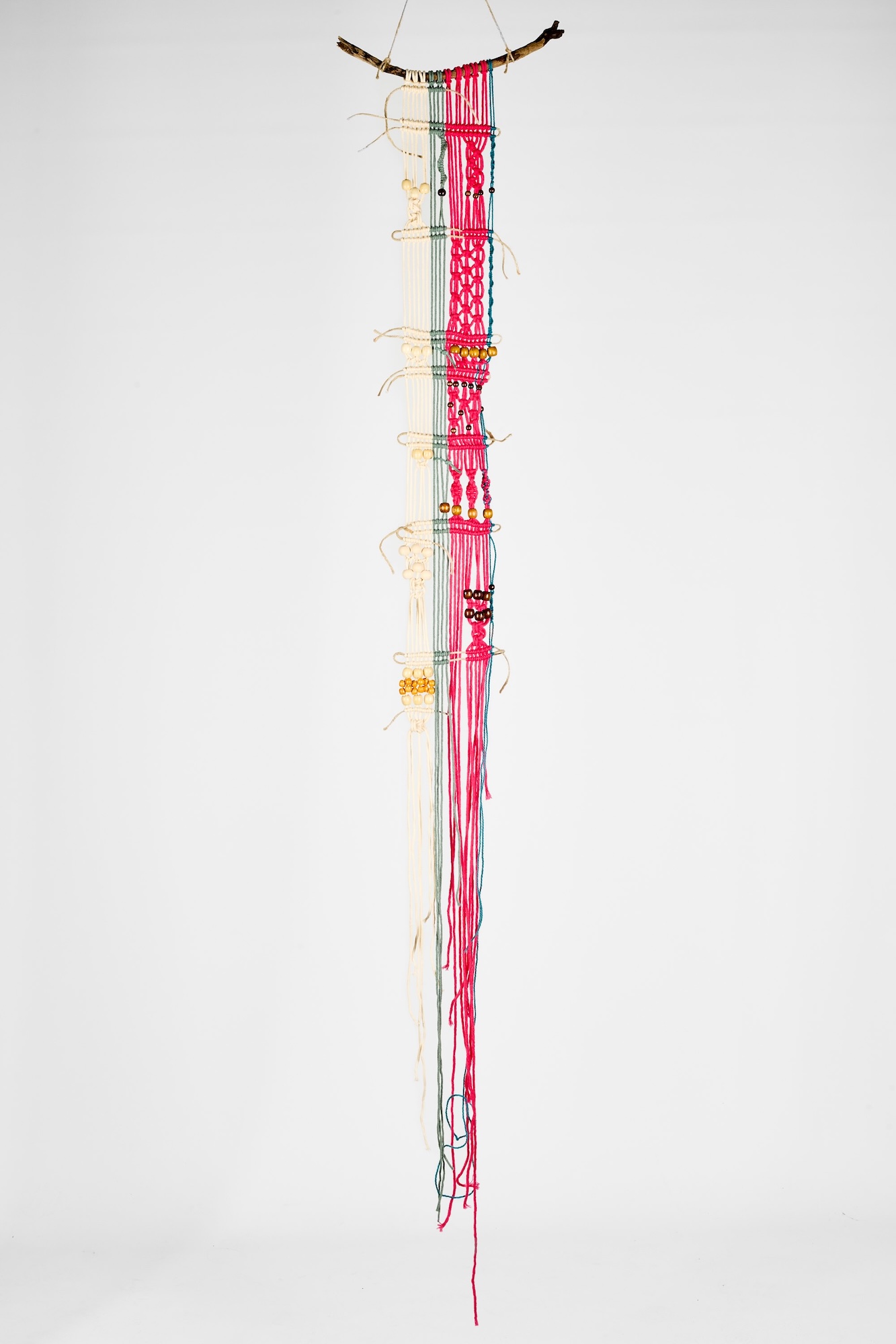 A long hanging wall sculpture of intertwined pink and cream cords, suspended from a curved branch against a plain white background.