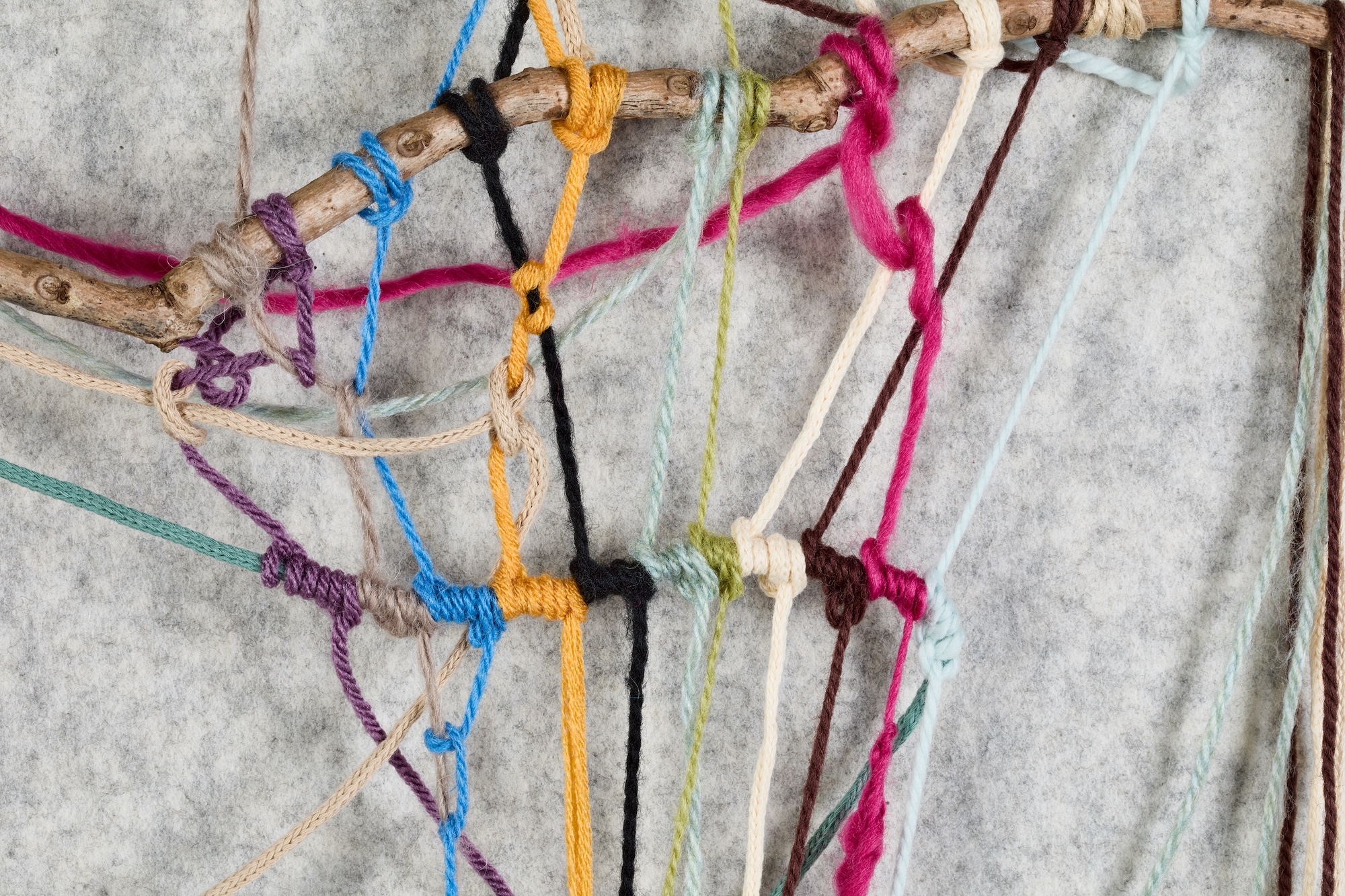 A close-up of multicoloured yarn threads tied around a branch against a rough grey background.