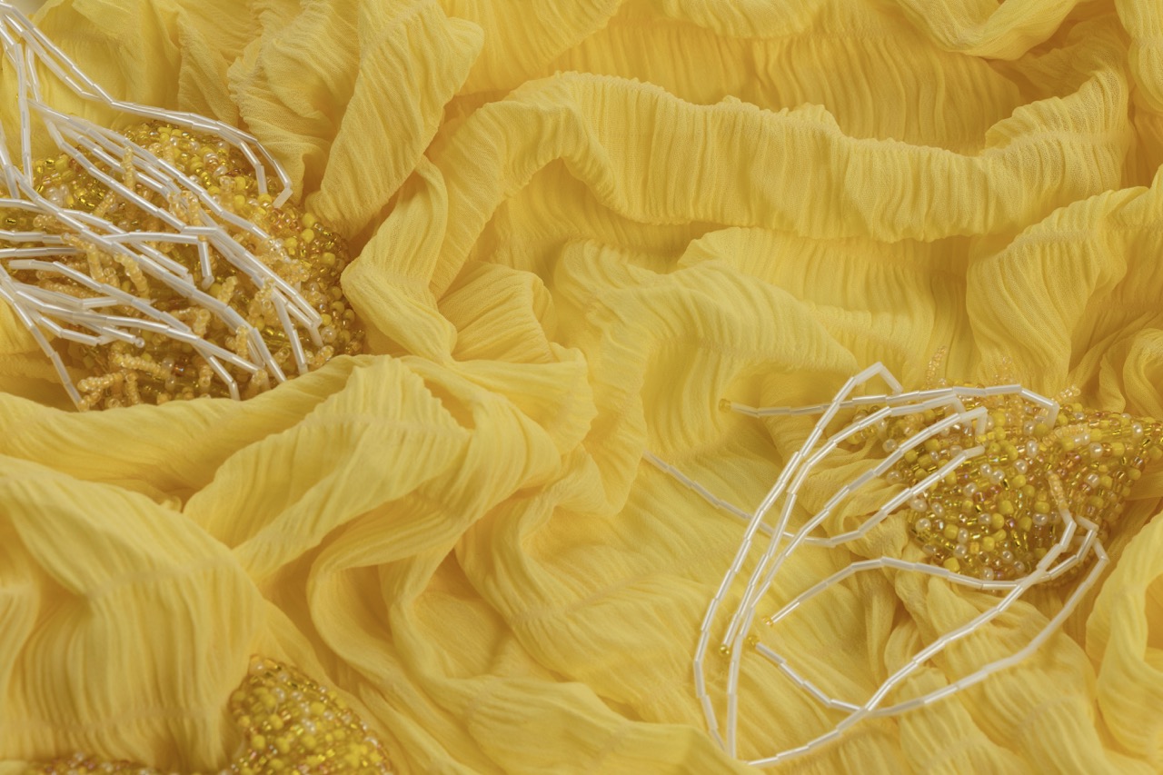 A close-up view of a bright yellow, textured fabric with folds and pleats. Beaded embellishments in yellow and white thread are scattered across the fabric, adding detail and pattern.