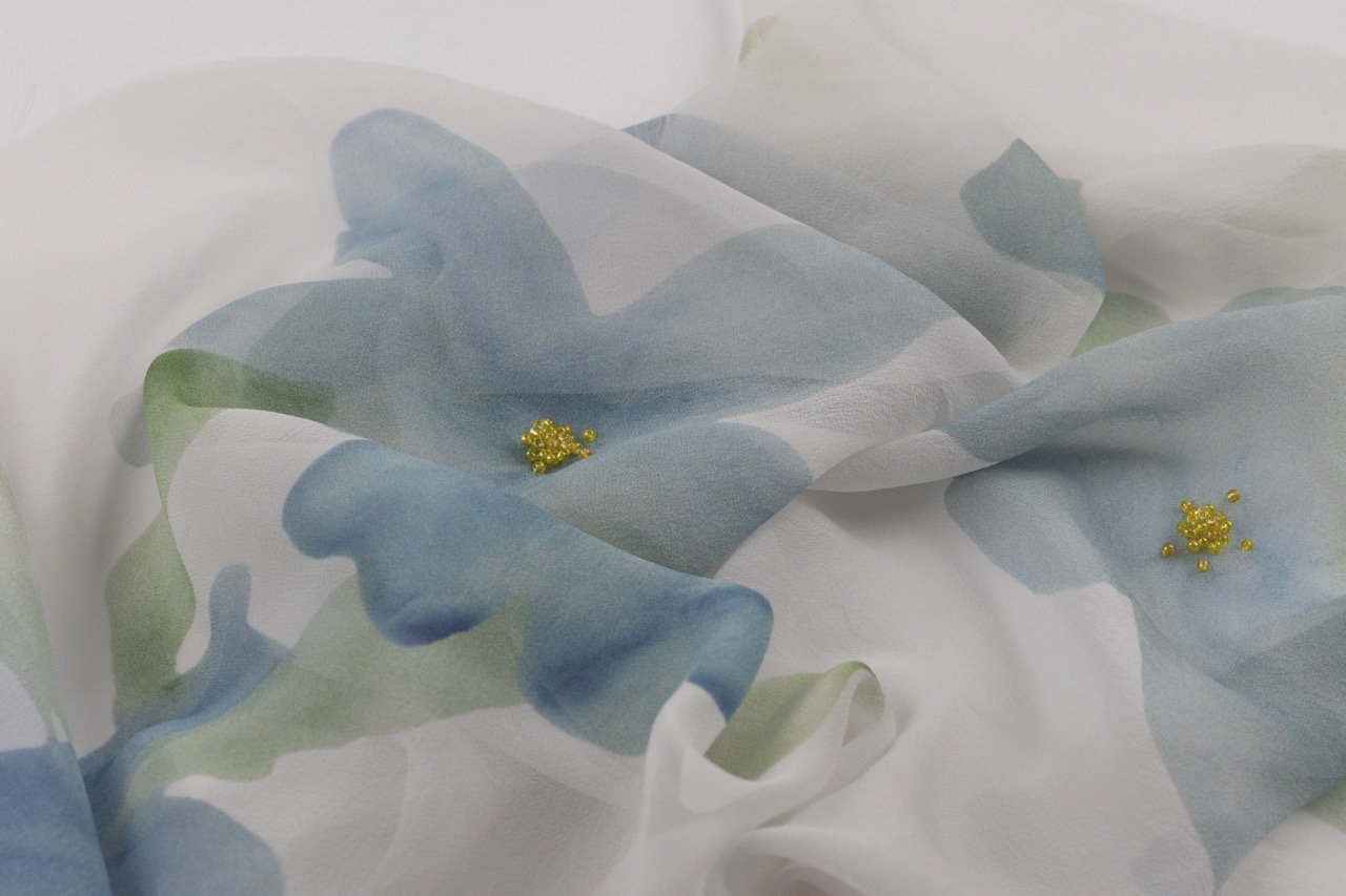 A sheer fabric featuring a floral pattern with large blue petals, accented by small yellow beads scattered across the surface. The delicate material is softly draped, allowing the light to pass through.