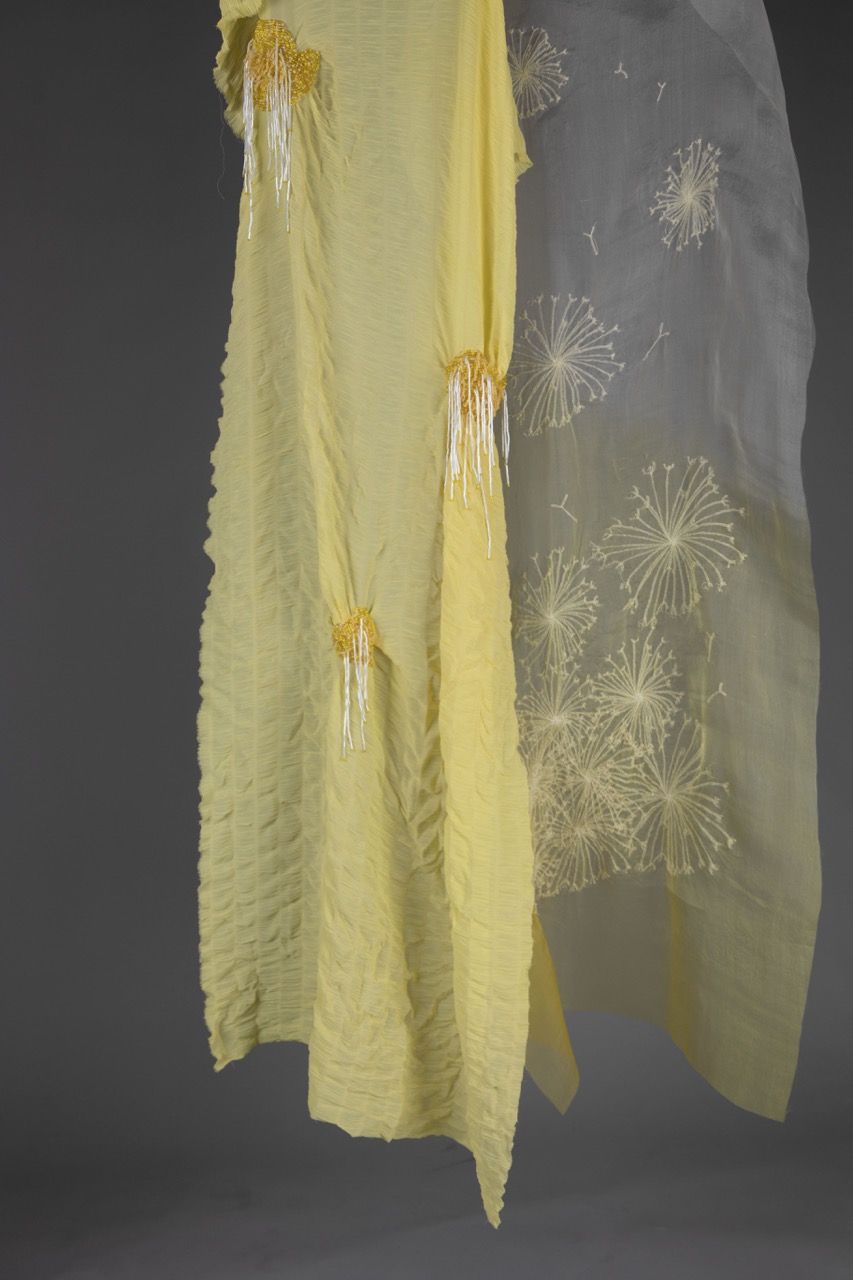 A long yellow garment made of light fabric features textured sections and intricate yellow embroidery at the top. Beside it hangs a sheer, grey fabric adorned with white firework-like patterns, both suspended against a dark background.