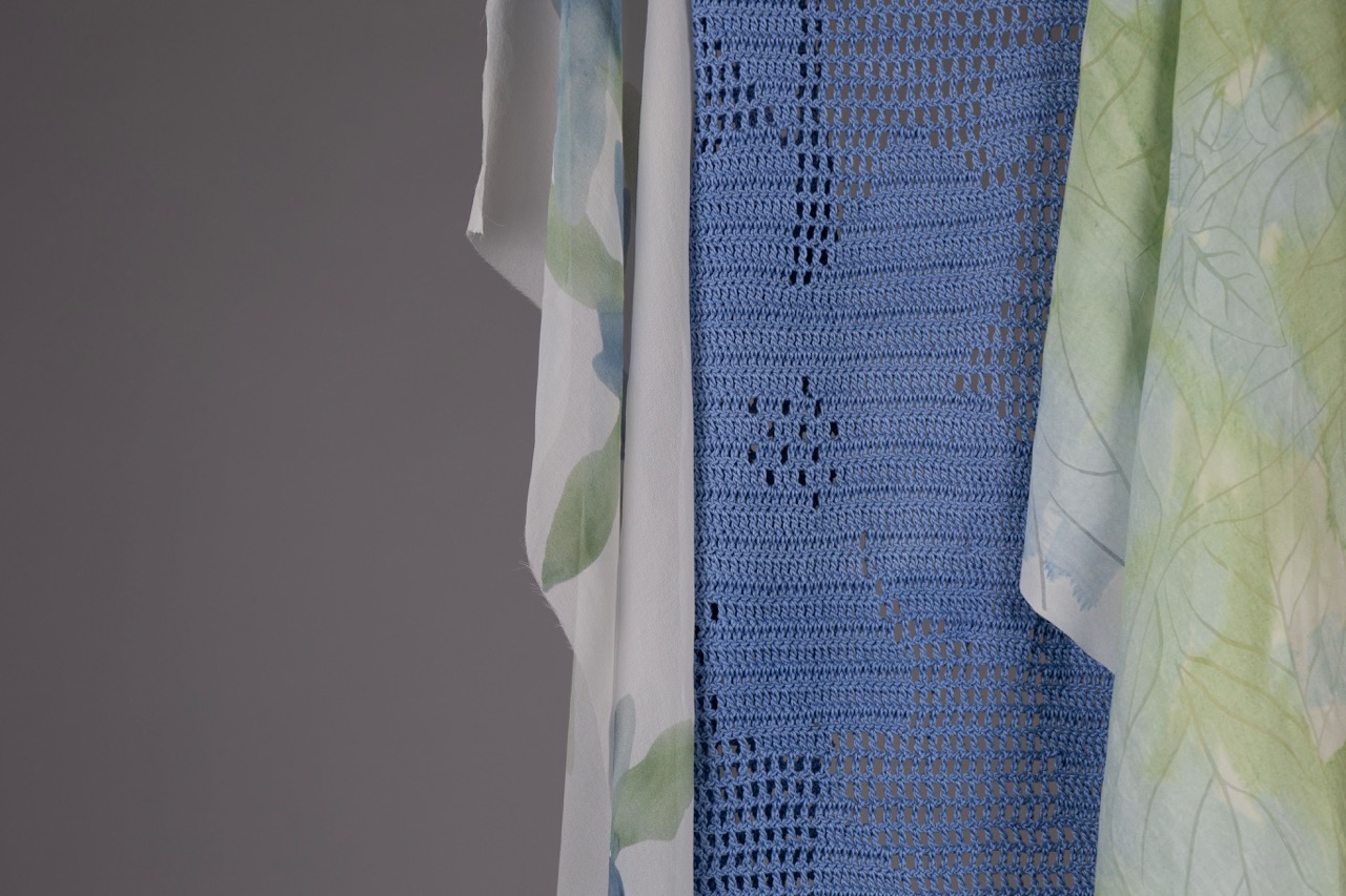 A collection of three pieces of fabric hanging side by side. The fabrics include a white one with green leaves, a blue crocheted piece, and a light green patterned fabric, against a grey background.