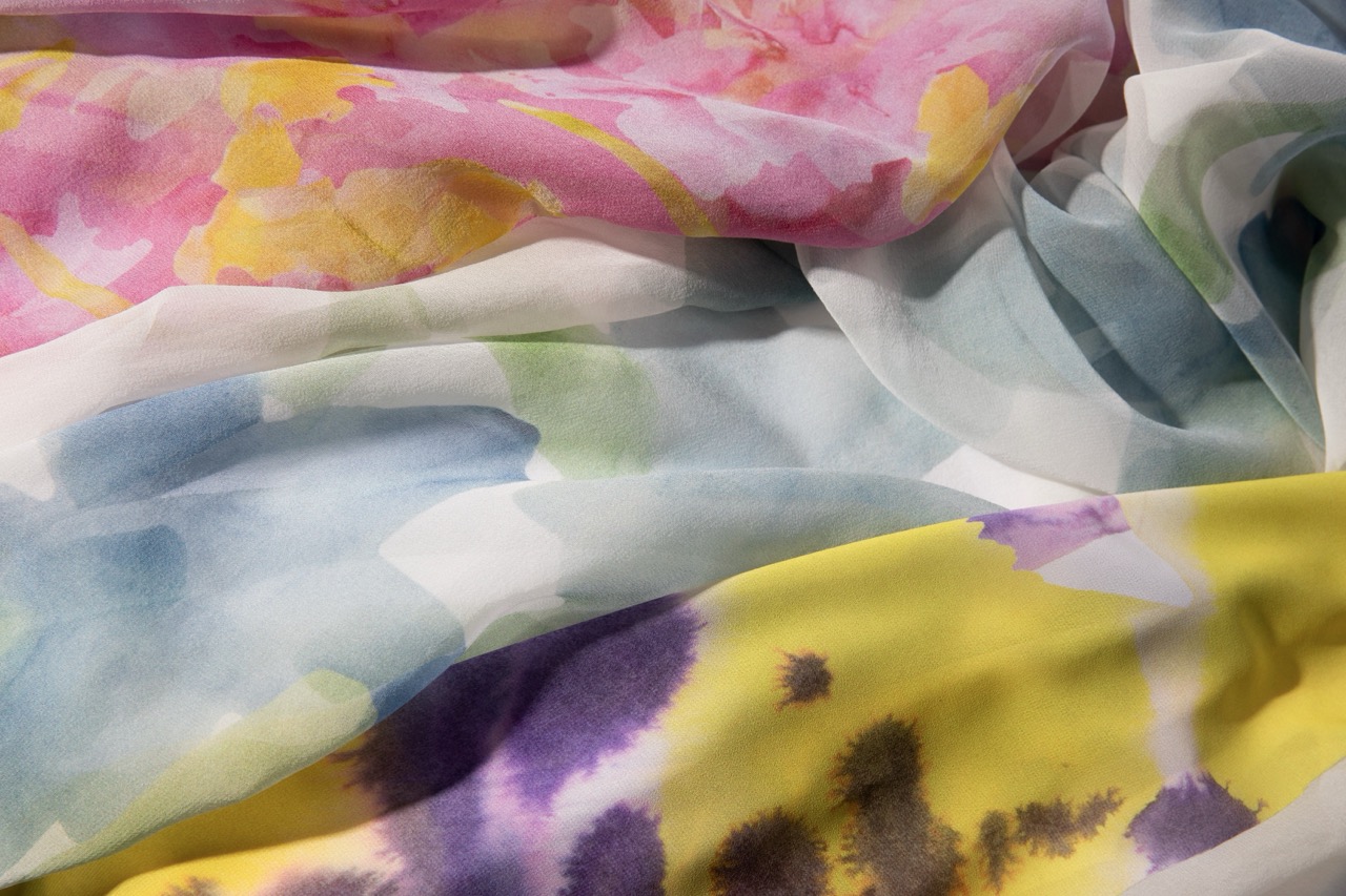 A close-up of lightweight, sheer fabric featuring a blend of pastel colours, including pink, yellow, and blue with floral patterns. The fabric appears soft and flowing, with vivid hues blending softly into one another.