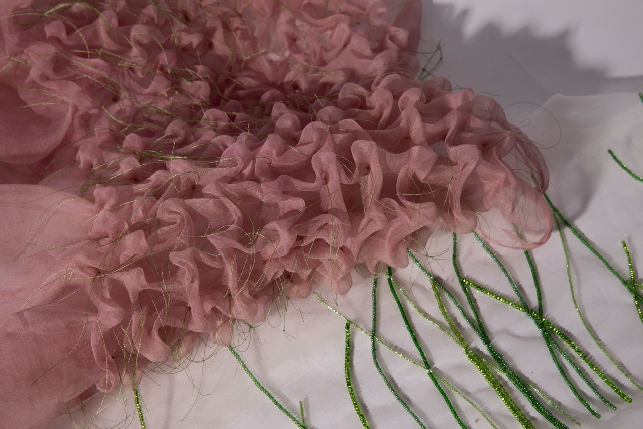 A close-up of pink sheer fabric featuring a ruffled texture, surrounded by green decorative strands. The fabric is layered in a wavy pattern, creating a soft and delicate appearance.