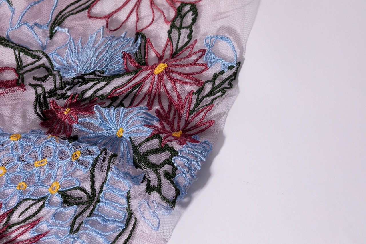 A piece of fabric featuring intricate embroidery with a variety of flowers in red, blue, and yellow hues. The design includes detailed floral patterns against a soft background, highlighting the fine stitching and artistry involved.
