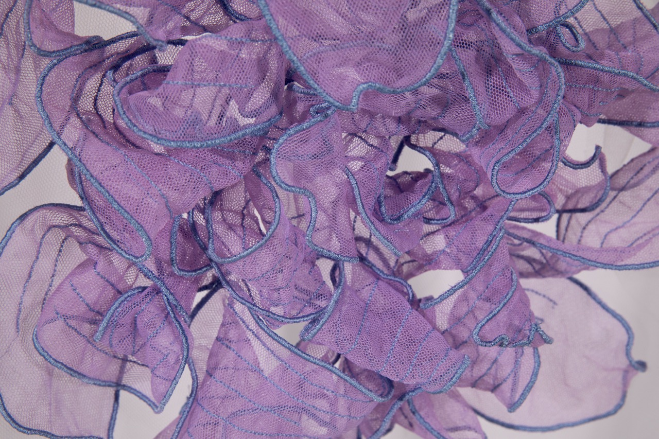 A close-up view of a tangled arrangement of sheer purple fabric with blue stitching along the edges. The fabric has a lightweight, airy quality and forms flowing, wavy shapes that create an intricate design.