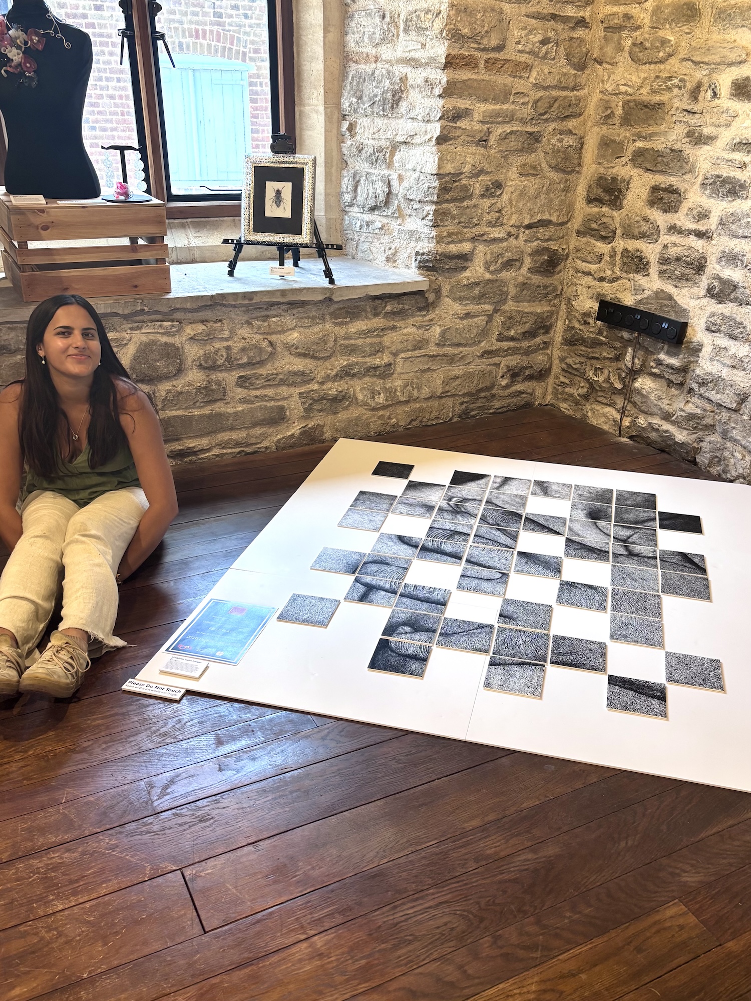 M Iyengar sitting cross-legged on a wooden floor beside a large black and white mosaic artwork. The artwork has a checkerboard pattern and is placed against a stone wall near a window.