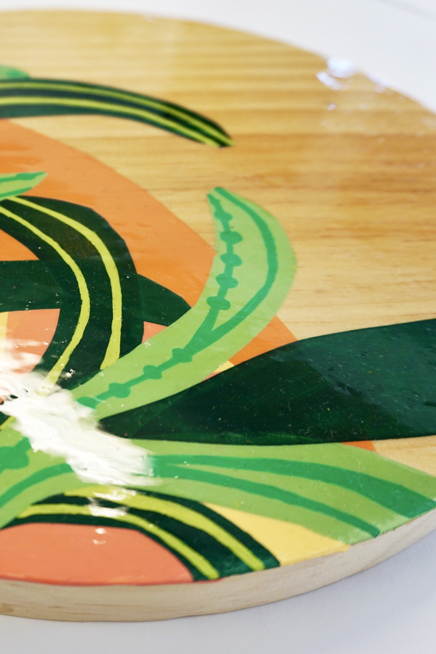 A circular wooden board is adorned with a colourful design featuring green leaves and orange shapes. The glossy surface reflects light, enhancing the vibrant colours of the artwork.