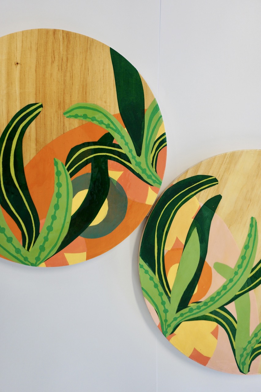 Two round wooden plates hanging on a white wall, featuring abstract designs with green leaves and colourful shapes in orange, pink, and grey tones. The plates are decorated with stylised illustrations of plants.