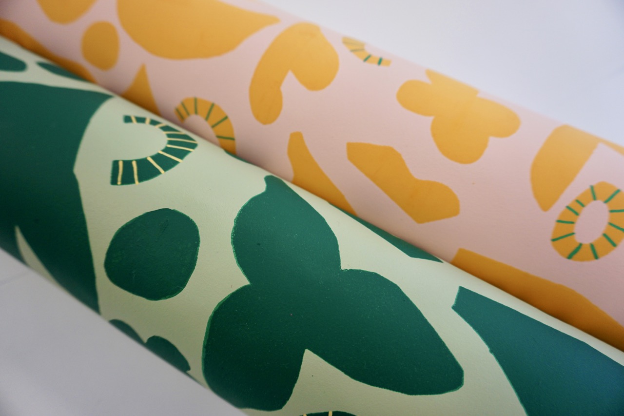 Two rolls of patterned wallpaper, one in pastel yellow featuring abstract shapes, and the other in green with overlapping leaf and floral motifs. Both rolls are placed on a light surface.