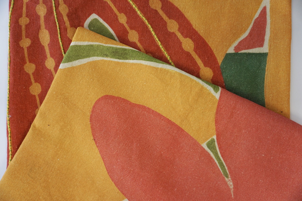 Two pieces of colourful fabric are overlapping. One has a bold design featuring a large red flower with green accents on an orange background, while the other has a pattern of yellow lines and dots on a mustard background.