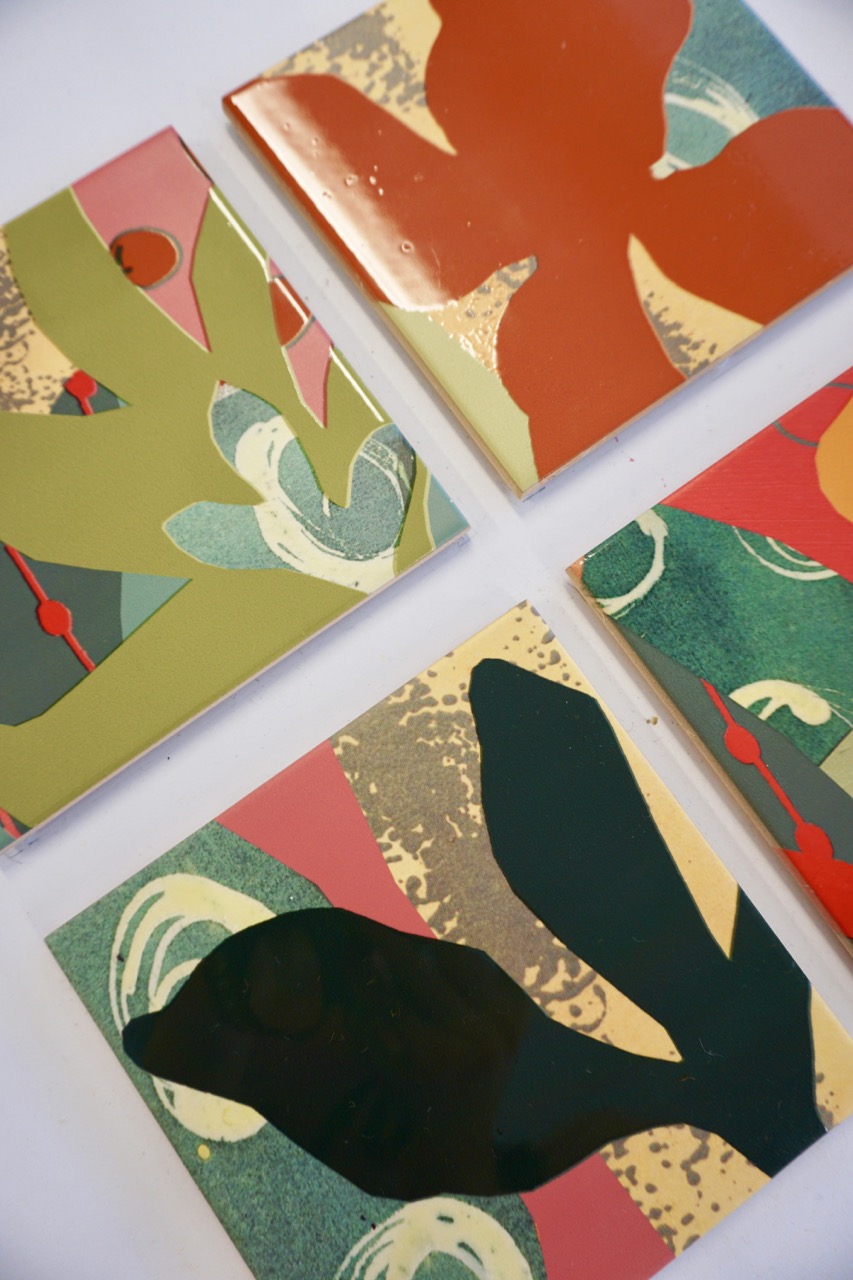 Four ceramic tiles arranged on a flat surface, showcasing various colourful designs. The tiles feature abstract floral shapes in red, green, and blue, set against textured backgrounds with contrasting patterns in yellow and beige.